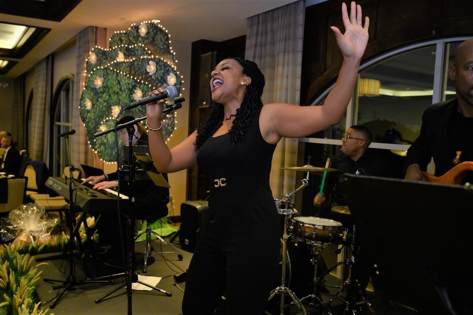 A woman with dark, curly hair singing passionately into a microphone while raising one arm during a musical performance. She is wearing a sleeveless black dress and appears to be in a decorated indoor venue with a Christmas-themed wall in the background, along with musicians playing instruments.