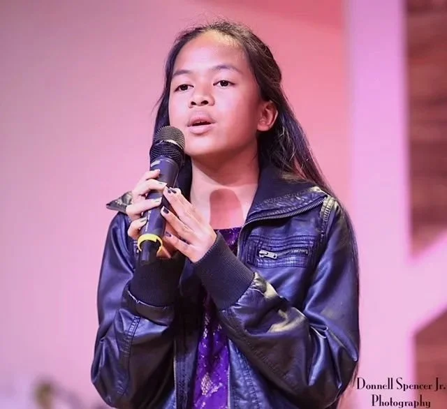 A young girl with long dark hair holding a microphone and speaking or singing on stage, wearing a black leather jacket and a purple shirt, with a plain pink background.