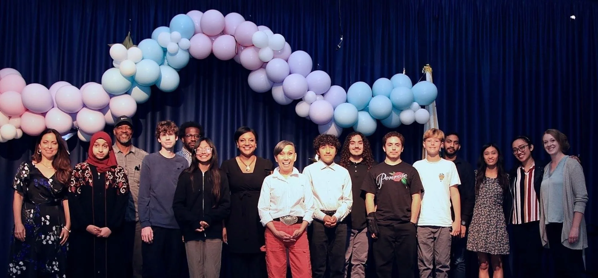 Group of diverse people standing on stage in front of blue curtain with colorful balloons overhead, celebrating an event or achievement.