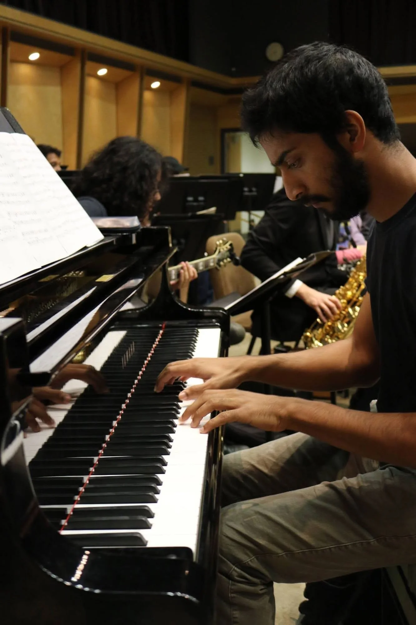 A young man playing the piano during an ensemble rehearsal, surrounded by musicians including a guitarist and a saxophonist, in a music studio with wooden paneling.