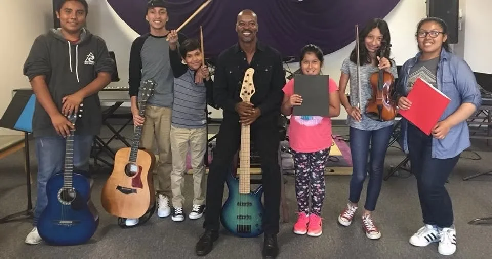 Group of seven children and one adult standing indoors, holding musical instruments including guitars, a violin, and a bass guitar. They are smiling and appear to be in a music classroom or rehearsal space.