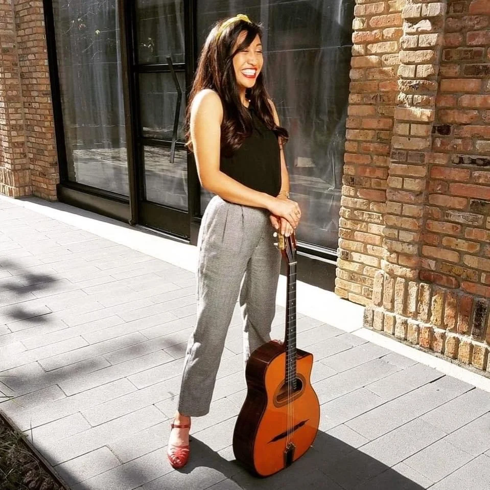 A young woman with long dark hair and a bright smile, holding an acoustic guitar on a sidewalk outside a brick building, wearing a black top, gray plaid pants, and red shoes.