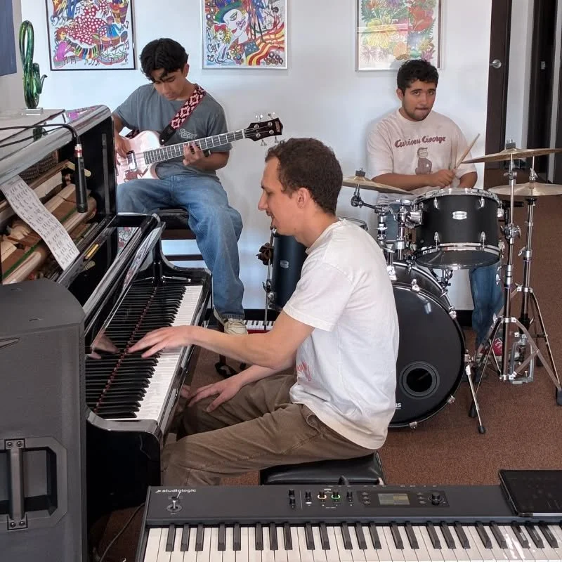 Group of three young men playing musical instruments indoors. One playing piano, one on drums, and one with guitar. Art posters on the wall behind them.