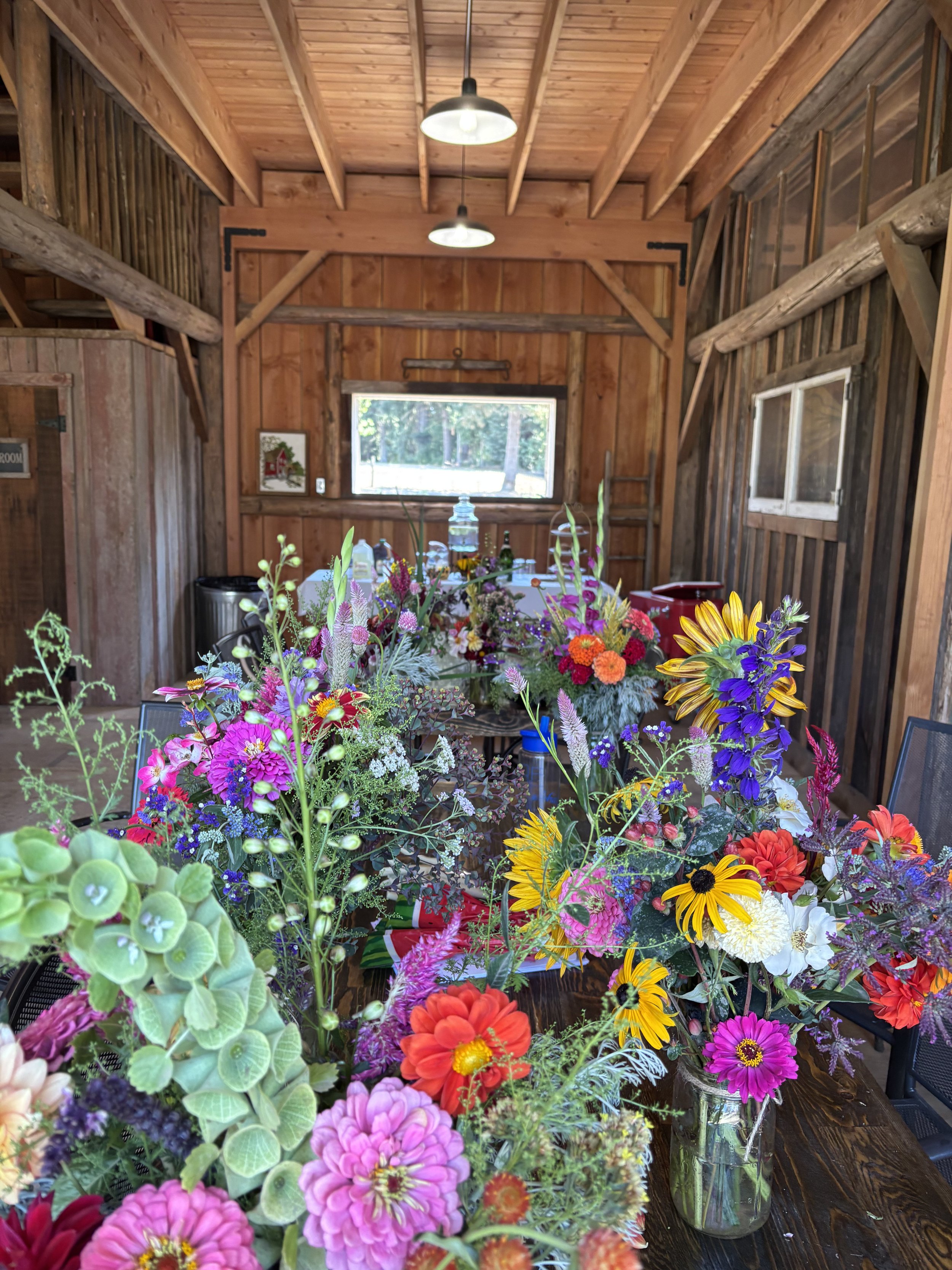 A rustic wooden room with a high ceiling and hanging lights. There is a large window letting in natural light and a table covered with colorful flower arrangements and vases.