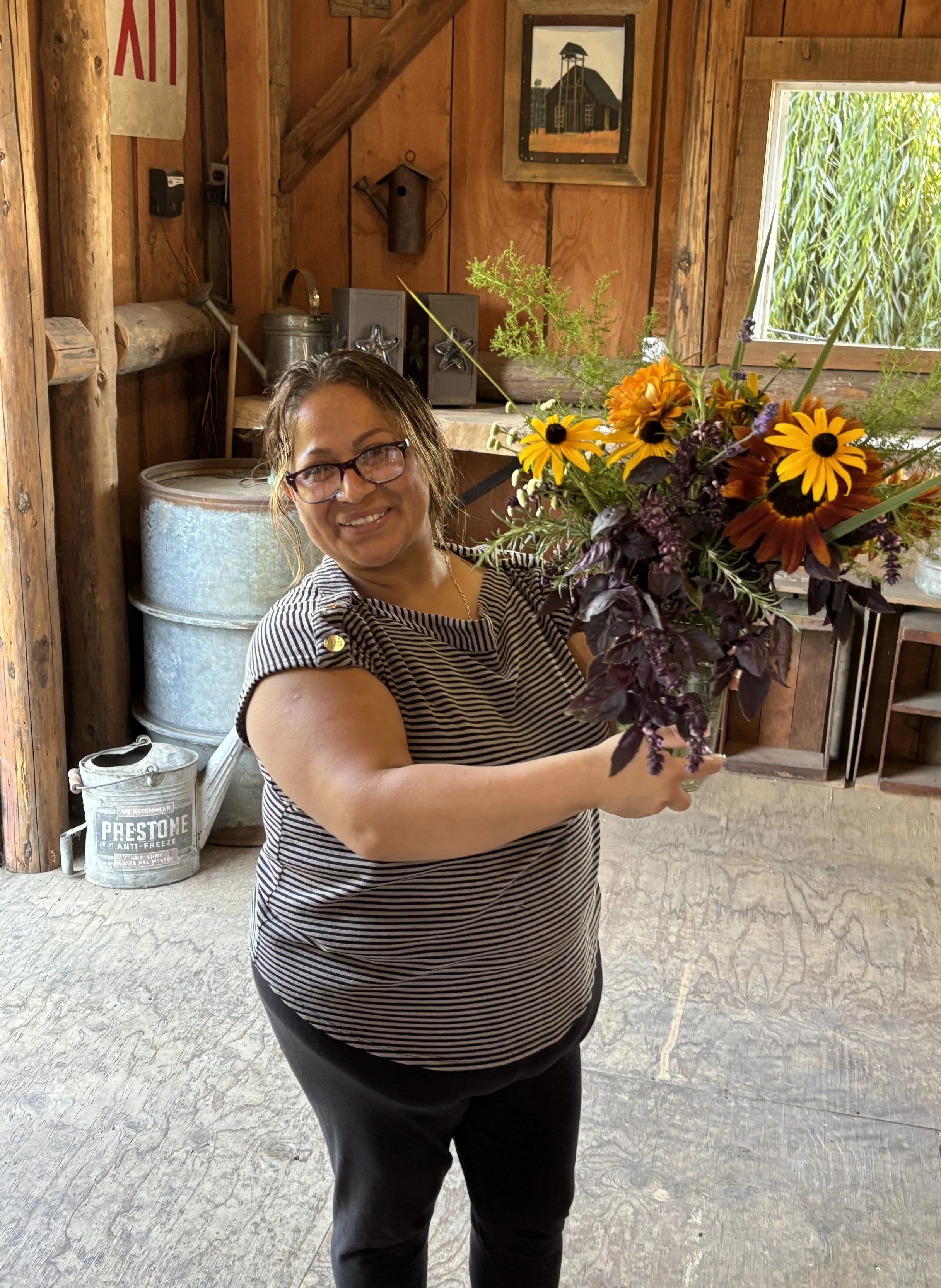 A woman wearing glasses and a striped shirt is holding a large bouquet of mixed flowers with yellow, orange, purple, and dark leaves in a rustic, wooden interior with various farm or barn decor.