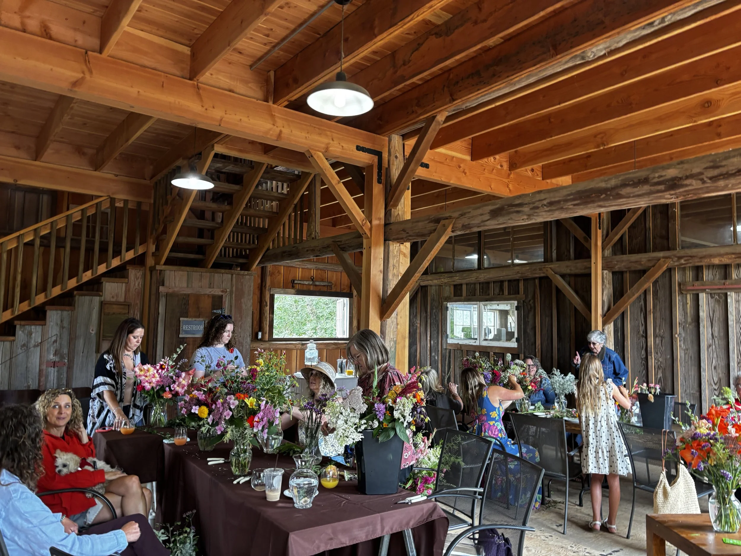People gathered indoors for a flower arranging workshop or event in a rustic wooden building with a high ceiling, wooden beams, and natural lighting. Participants are working with colorful flowers on a long table.