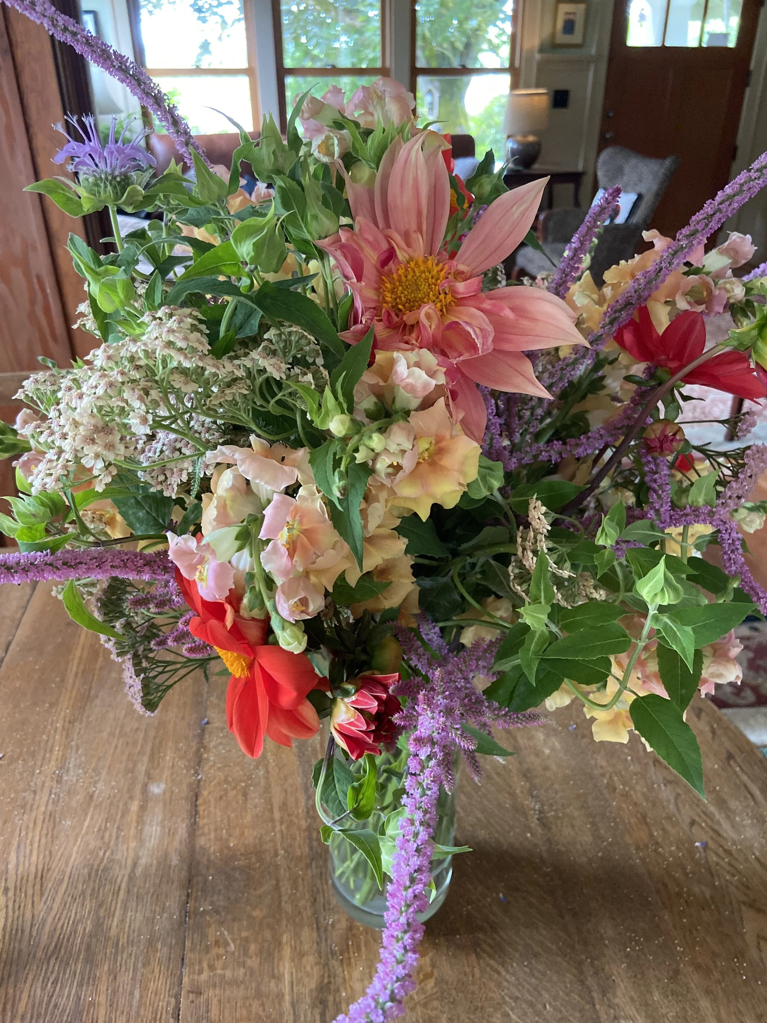 A colorful bouquet of flowers in a glass vase on a wooden table, with a background of windows and a living room.
