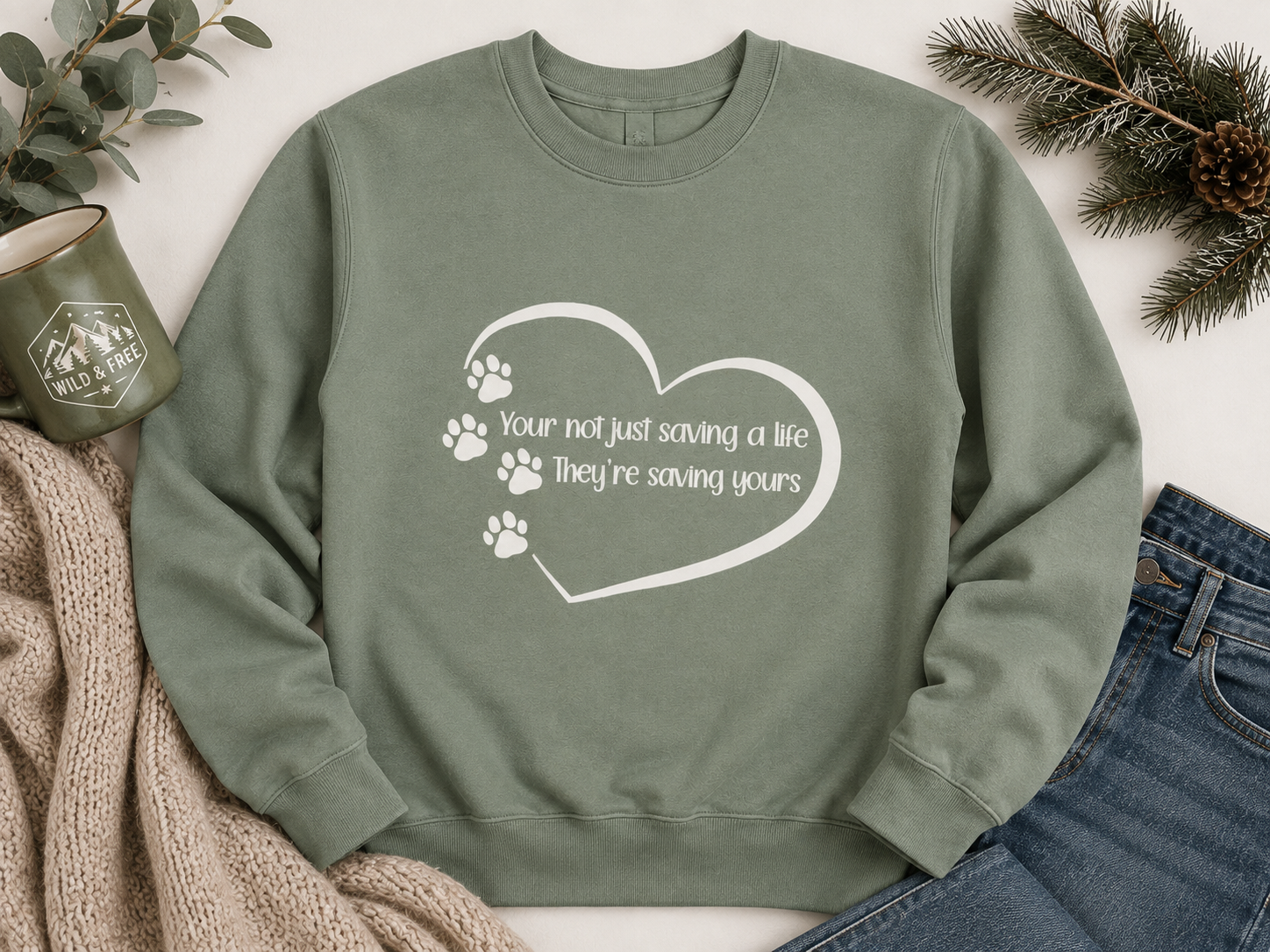 Green sweatshirt with white text inside a heart outline, surrounded by paw prints. The text reads: "You're not just saving a life. They're saving yours." The sweatshirt is laid out on a flat surface with a mug, pine branches, and denim jeans nearby.