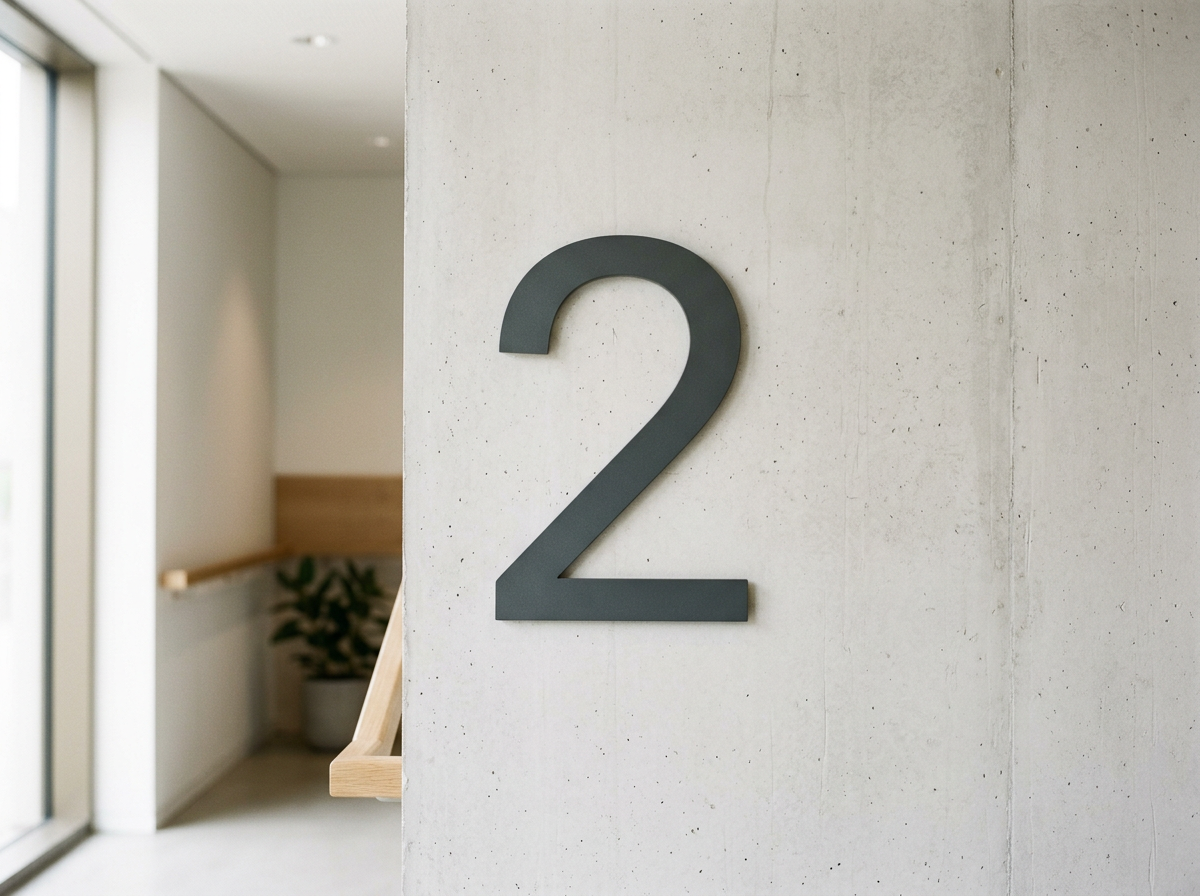 Interior of a building with a large black number '2' on a concrete wall, a staircase with a wooden handrail, and a potted plant in the background.