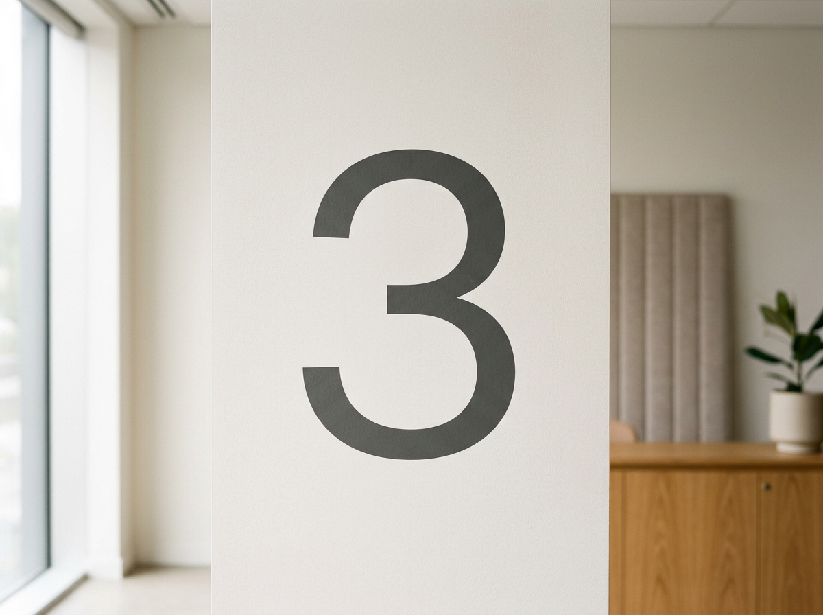 A large, black number three printed on a white wall, part of a room with a wooden piece of furniture and a potted plant visible in the background.