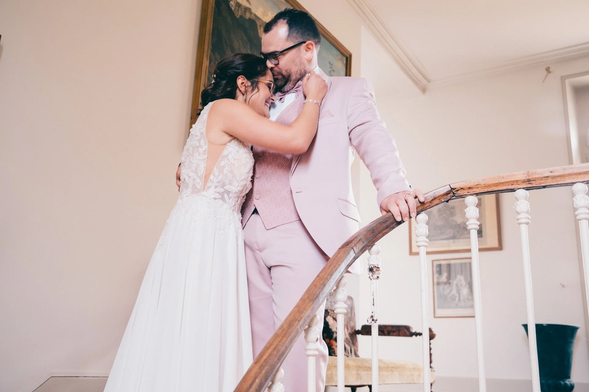 Un couple habillé en mariage, l'homme en costume rose et la femme en robe blanche, se tenant proche sur un escalier intérieur, potentiellement lors d'un mariage.