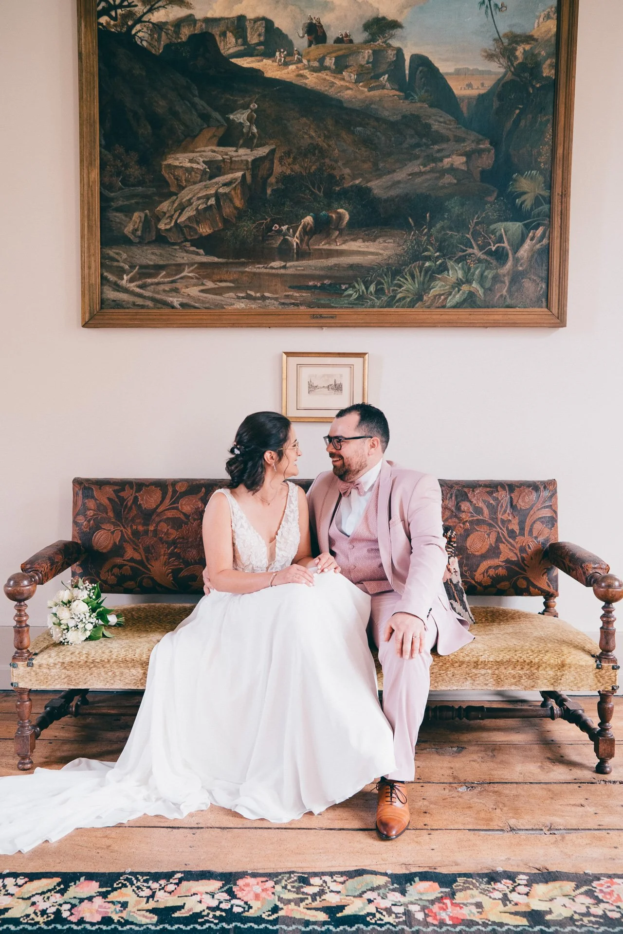 Un couple marié assis sur un canapé en bois et tissu tissé, se regardant et souriant. La femme porte une robe de mariage blanche et le homme un costume rose pâle avec une cravate. Il y a un bouquet de fleurs blanches à côté d'eux. Un tableau représen