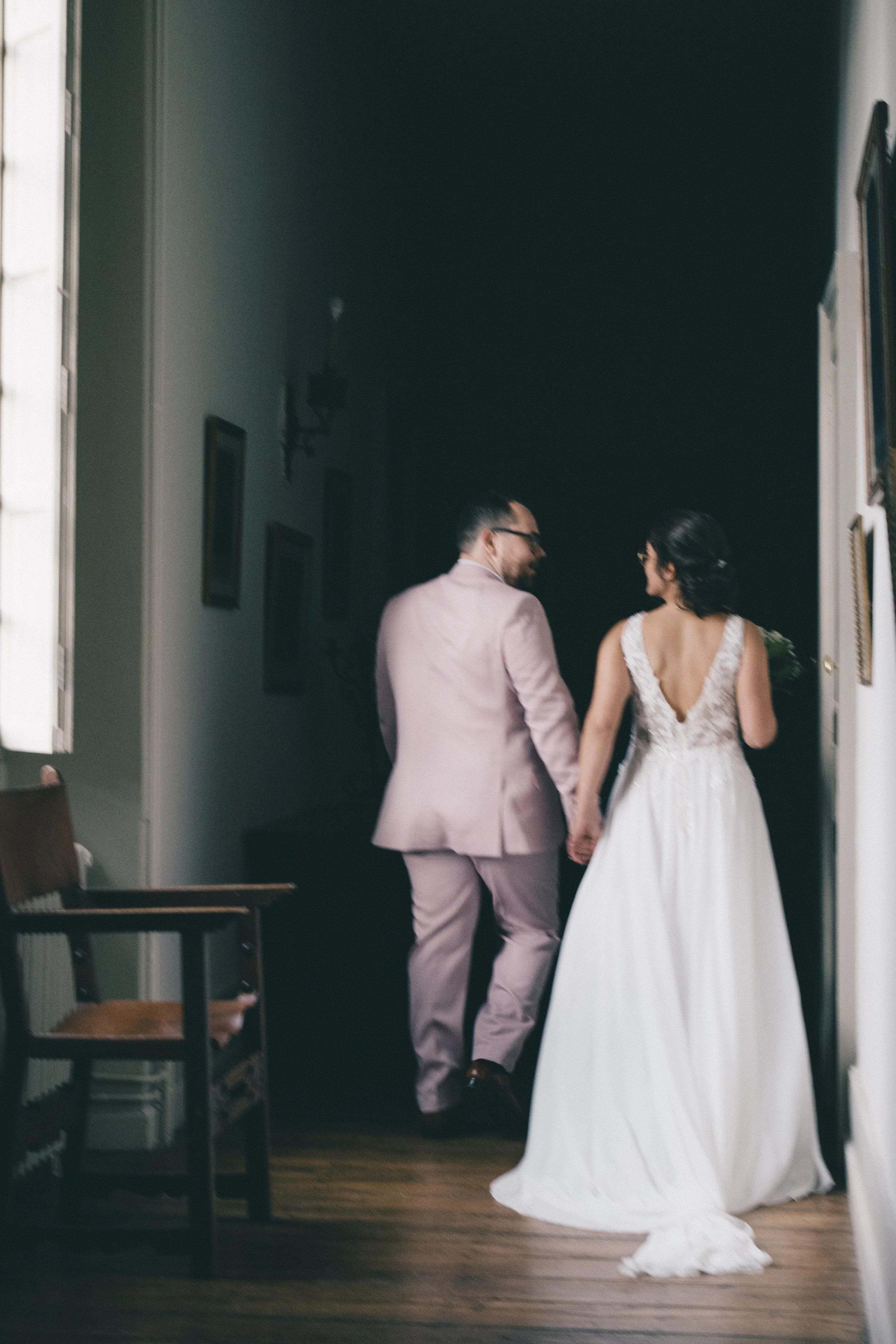 Un couple de mariés marche dans une pièce, vue de derrière. Le marié porte un costume rose, la mariée une robe de mariage blanche. Ils tiennent la main, discutent et semblent heureux.