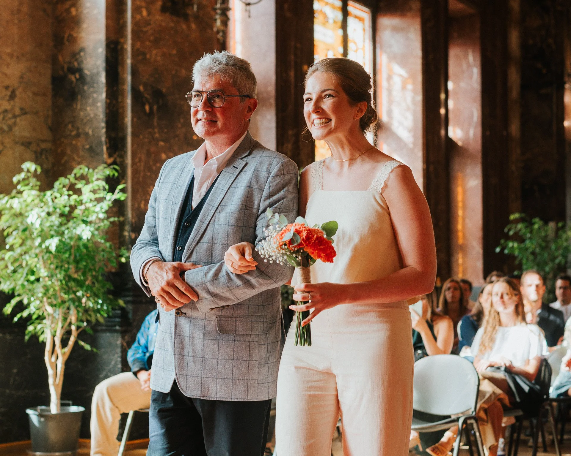 Un homme âgé en costume gris et une jeune femme en robe de mariage tiennent la main lors d'une cérémonie de mariage dans une salle éclairée par la lumière naturelle, avec des invités assis en arrière-plan.
