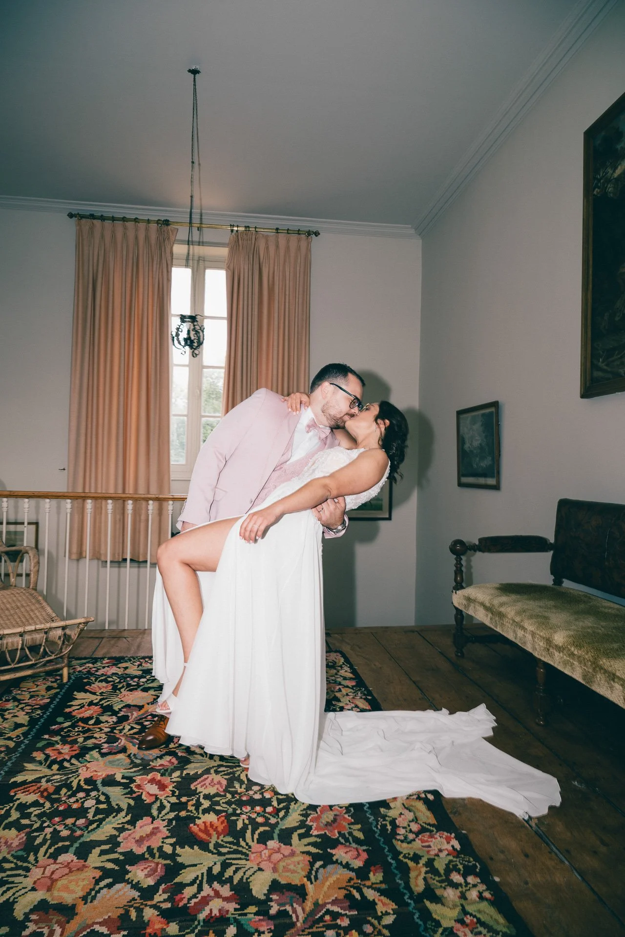 Un couple danse dans une pièce, la femme portant une robe blanche longue, l'homme en costume clair. Ils s'embrassent, le fond montre des fenêtres avec des rideaux beiges, des tableaux accrochés au mur et un tapis coloré au sol.