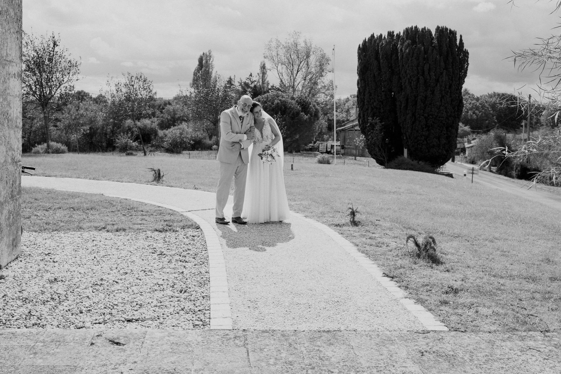 Un couple en costume de mariage marche main dans la main sur un chemin dans un parc, entouré d'arbres et d'une végétation arbustive.