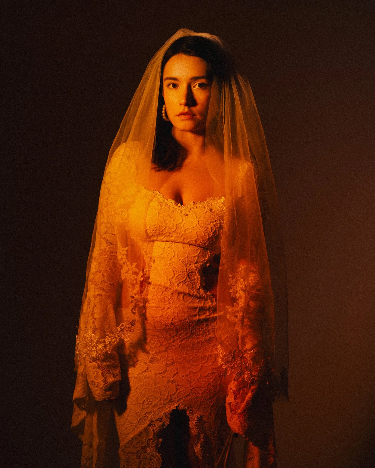Une femme portant une robe en dentelle orange et un voile assorti, éclairée par une lumière chaude, contre un fond sombre.