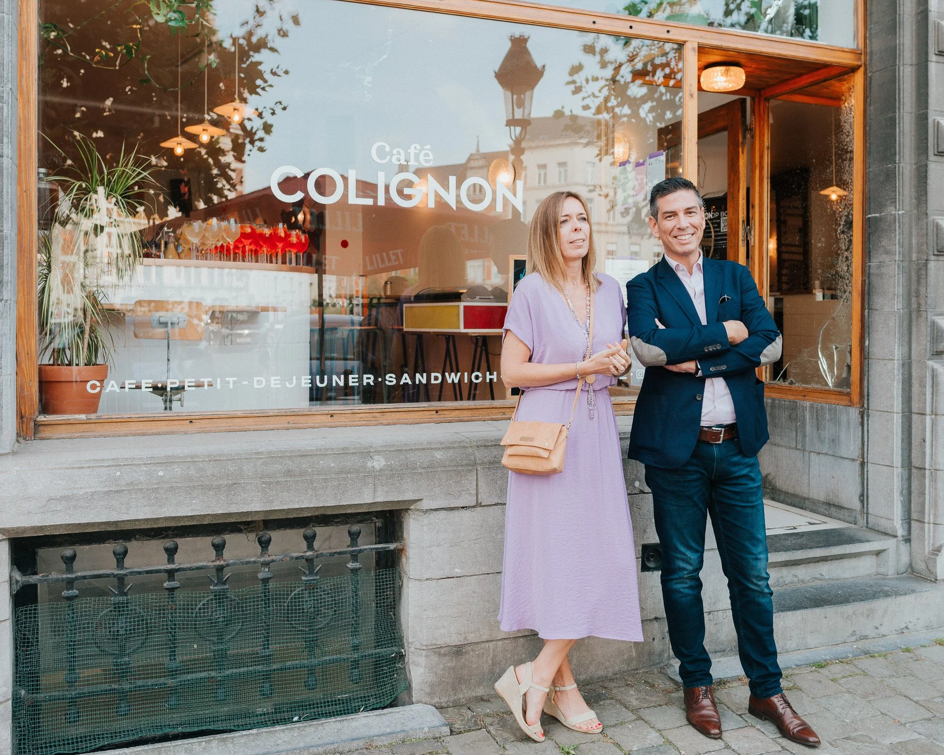 Deux personnes en costume, une femme en robe lilas et un homme en costume sombre, posent devant une fenêtre d'un café appelé Café Colignon à Paris, avec des décorations intérieures et un lampadaire reflétés dans la vitre.
