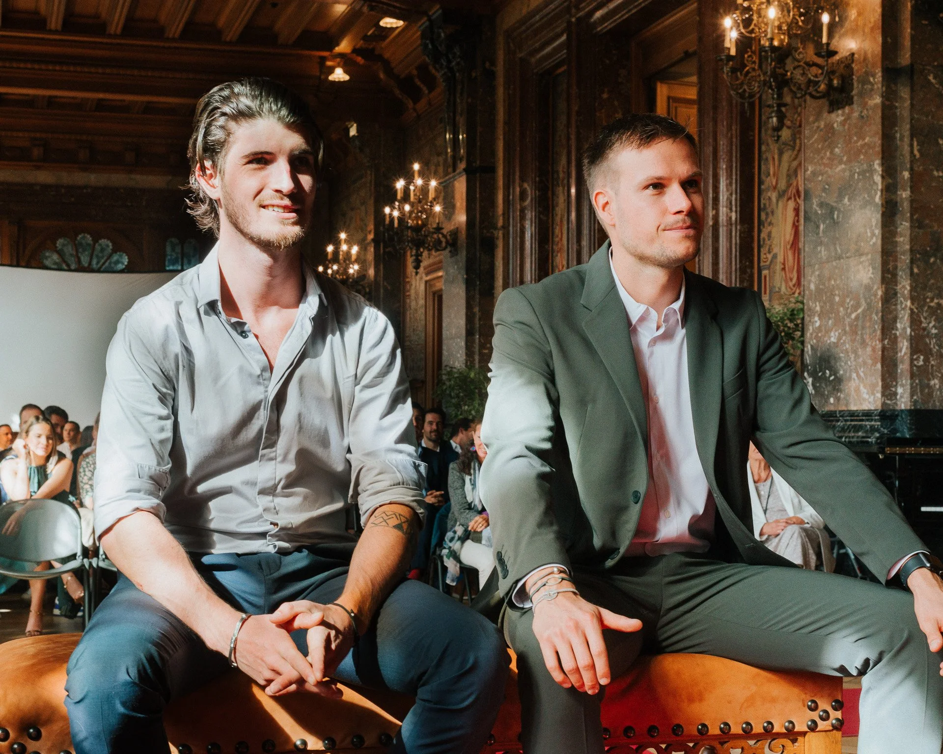 Deux jeunes hommes assis côte à côte lors d'un événement dans une salle élégante avec chandeliers. L'un porte une chemise gris clair, l'autre un costume gris avec une chemise blanche.