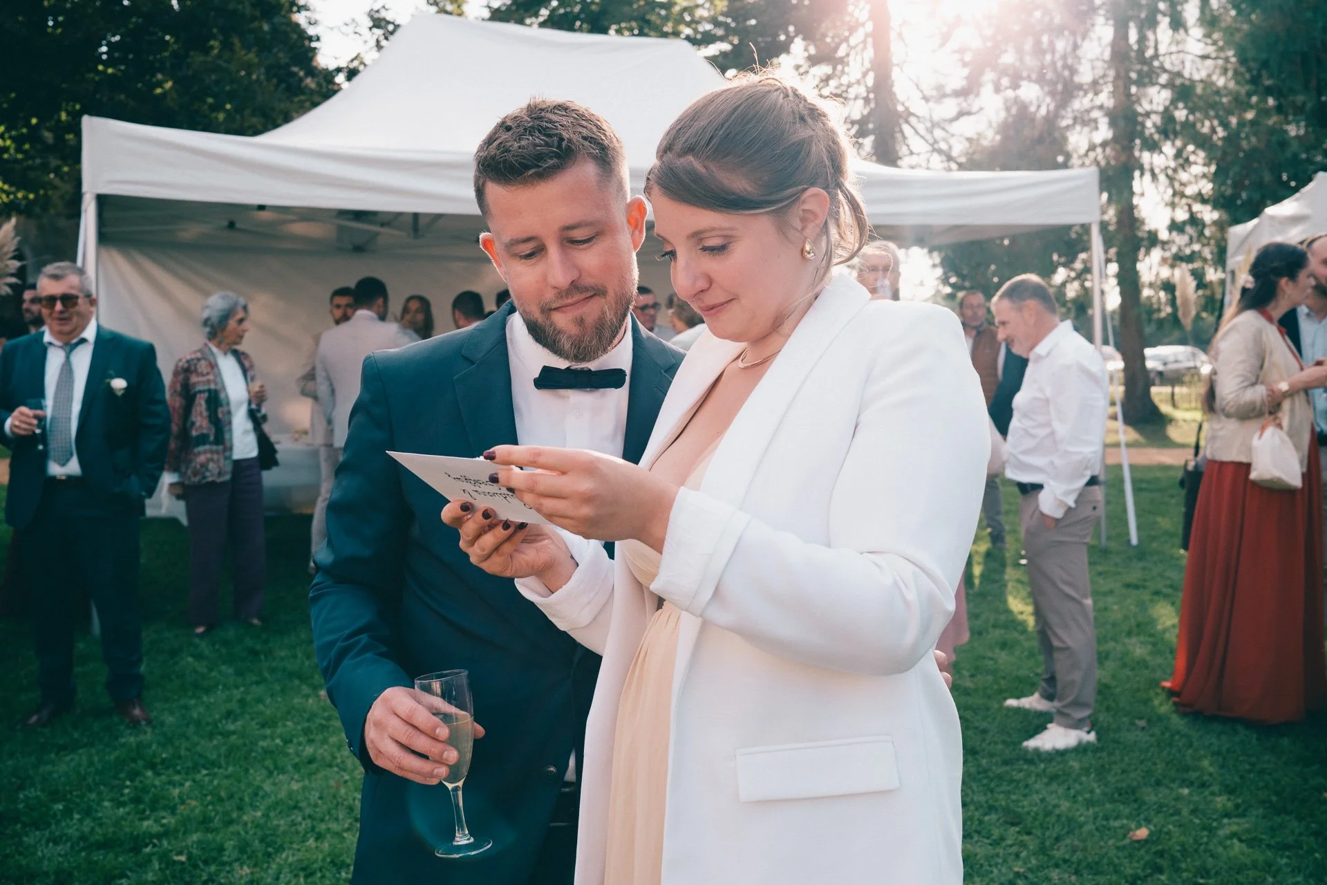 Un couple habillé formellement lors d'une réception en plein air, avec d'autres personnes en arrière-plan près de tentes. La femme porte un blazer blanc et la manœuvre un téléphone, tandis que l'homme porte un costume sombre avec un nœud papillon. La