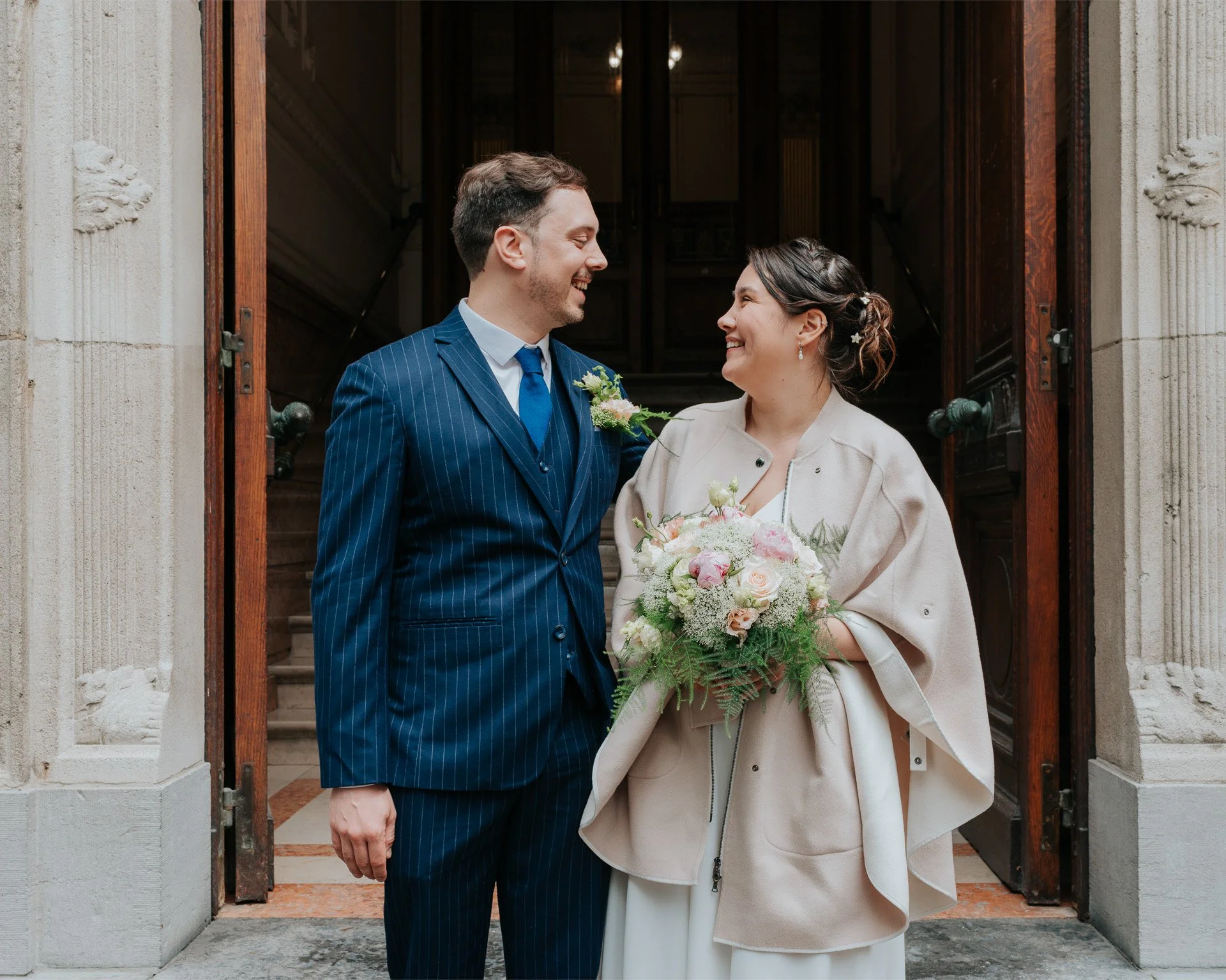 Un couple souriant, habillé pour une occasion spéciale, se tenant devant une porte en bois, la femme tenant un bouquet de fleurs.
