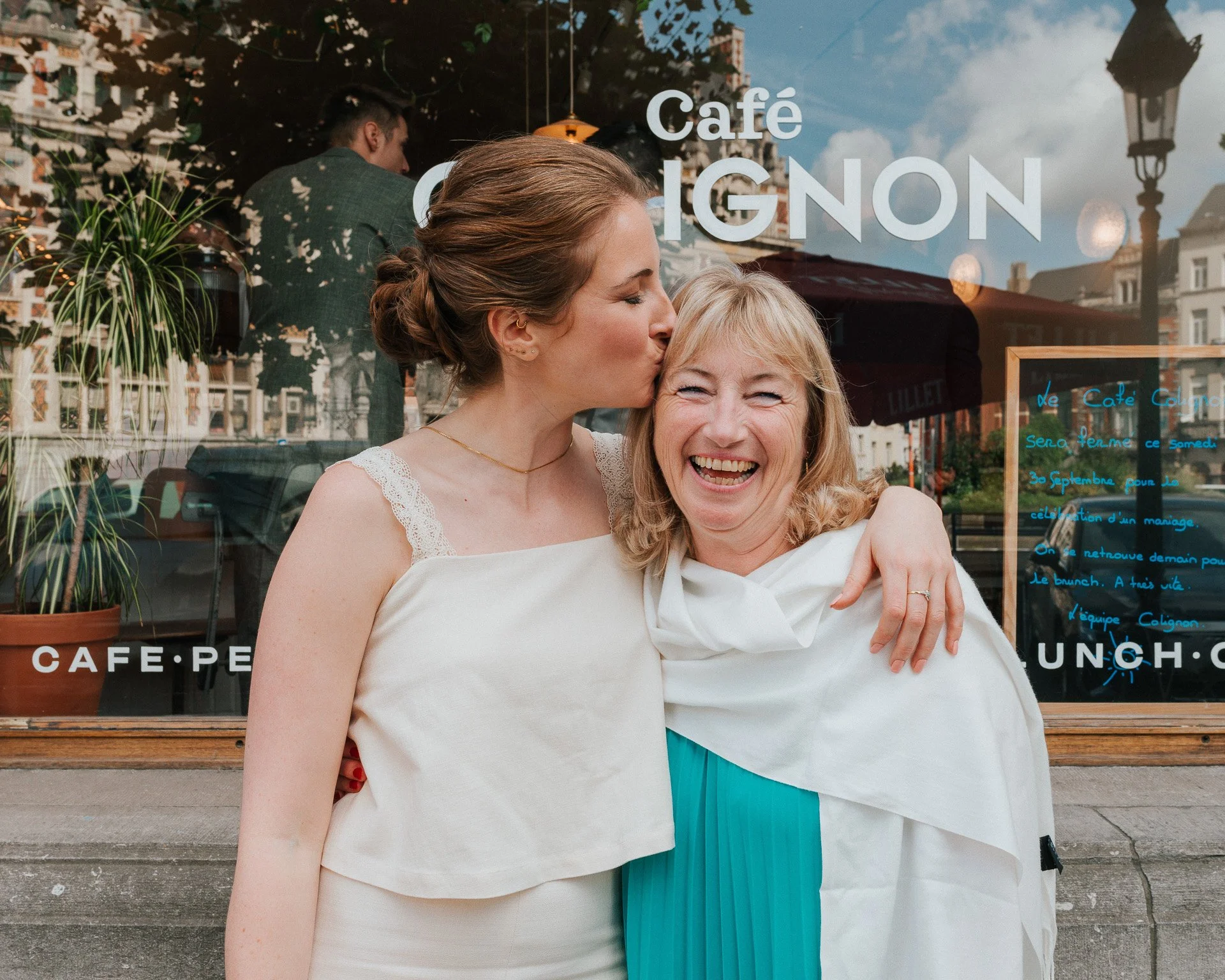Deux femmes, l'une avec les cheveux bruns attachés en chignon, donne un baiser sur le front à une femme plus âgée aux cheveux blonds, souriant.