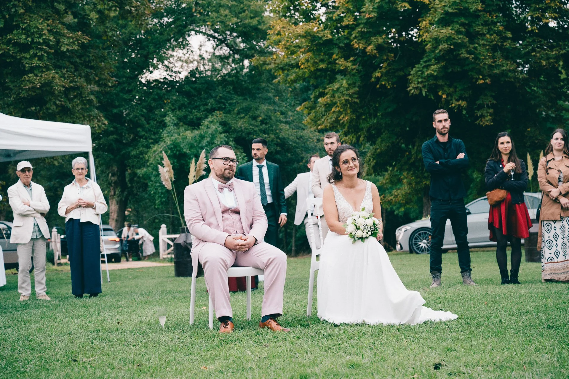 Un couple en vêtements de mariage assis dans un jardin lors d'une cérémonie. Autour d'eux, des invités regardent la célébration, dans un environnement vert avec des arbres en arrière-plan.