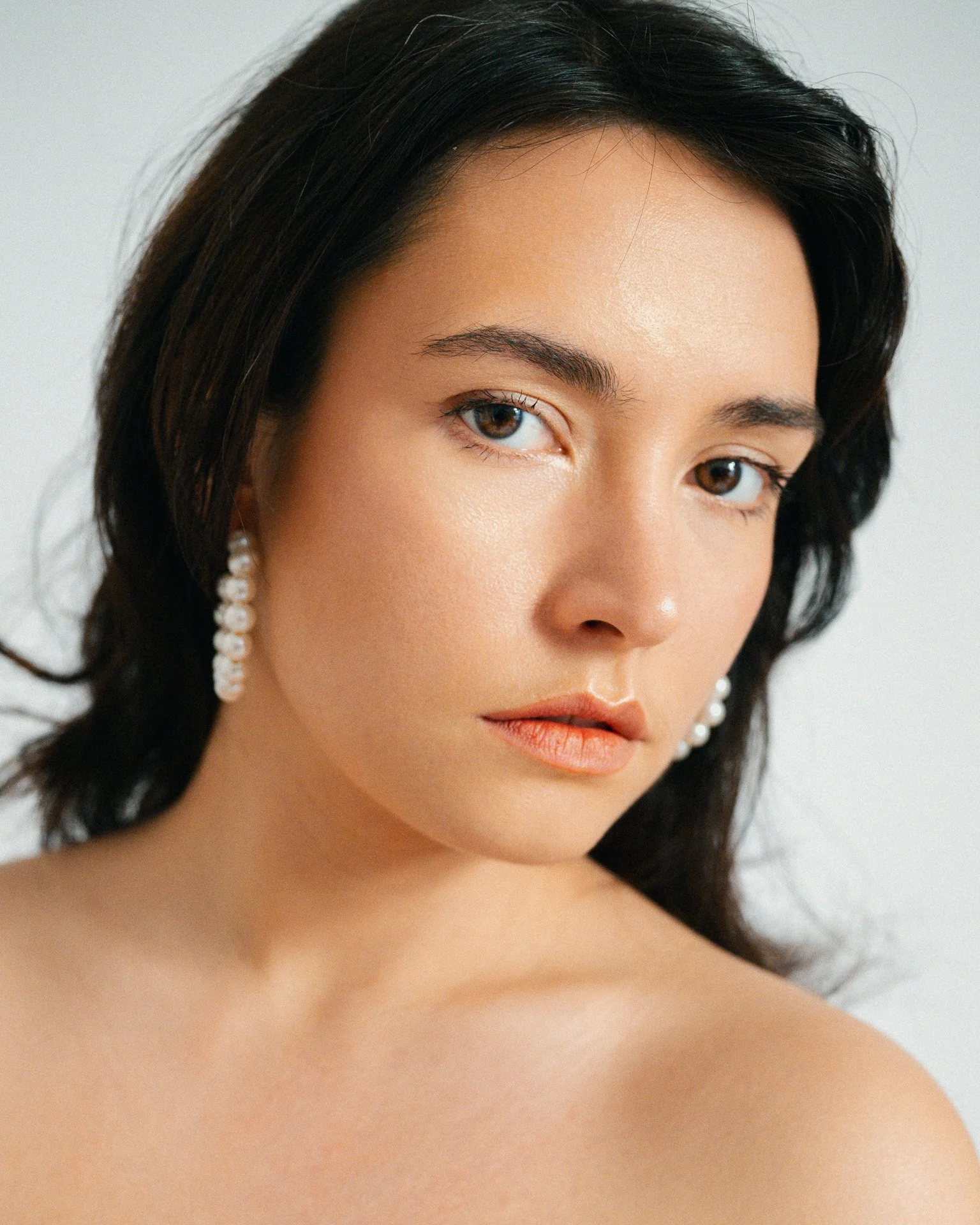 Close-up d'une femme avec des cheveux noirs, portant des boucles d'oreilles en perles, peau claire, maquillage léger, regardant vers la caméra.