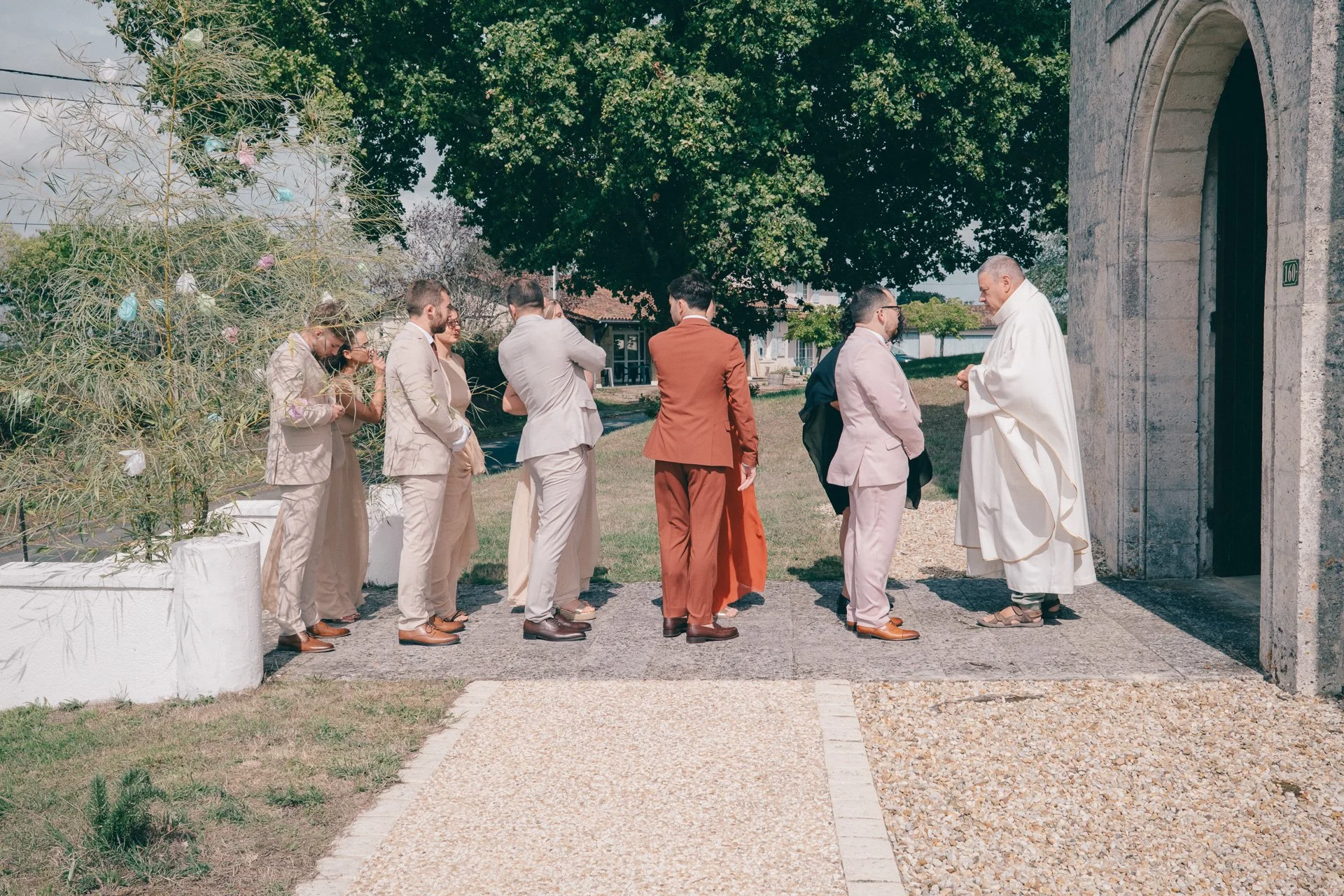 Un prêtre en robe blanche bénit un groupe de personnes habillées en costumes lors d'une cérémonie religieuse à l'extérieur, devant une église en pierre, avec des arbres en arrière-plan.