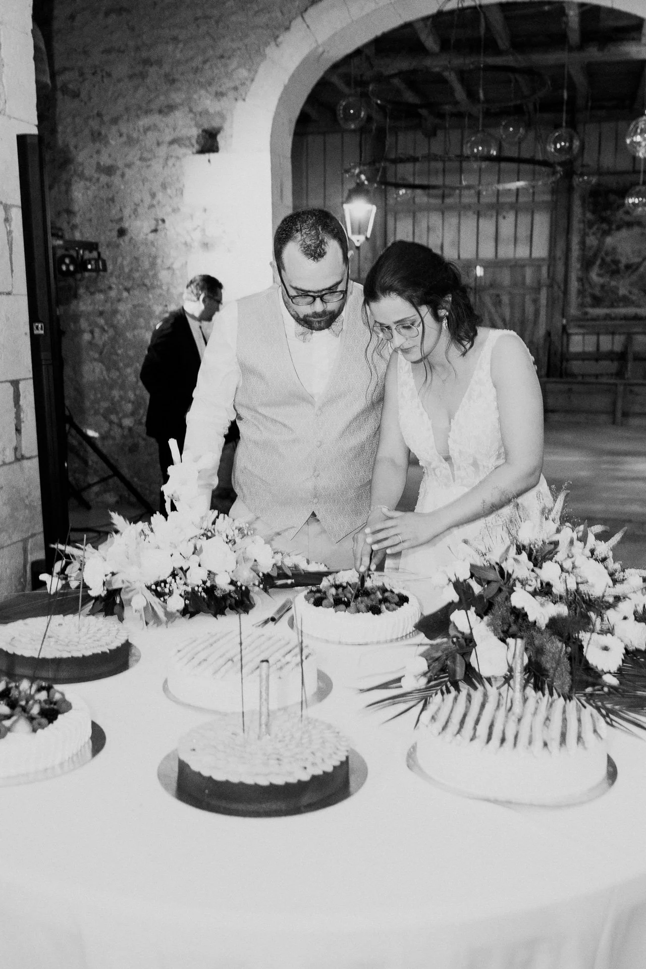Un couple en mariage coupe un gâteau de mariage décoré avec des fleurs, entouré de plusieurs autres gâteaux avec des fleurs, dans une salle rustique en bois lors d'une célébration nuptiale.