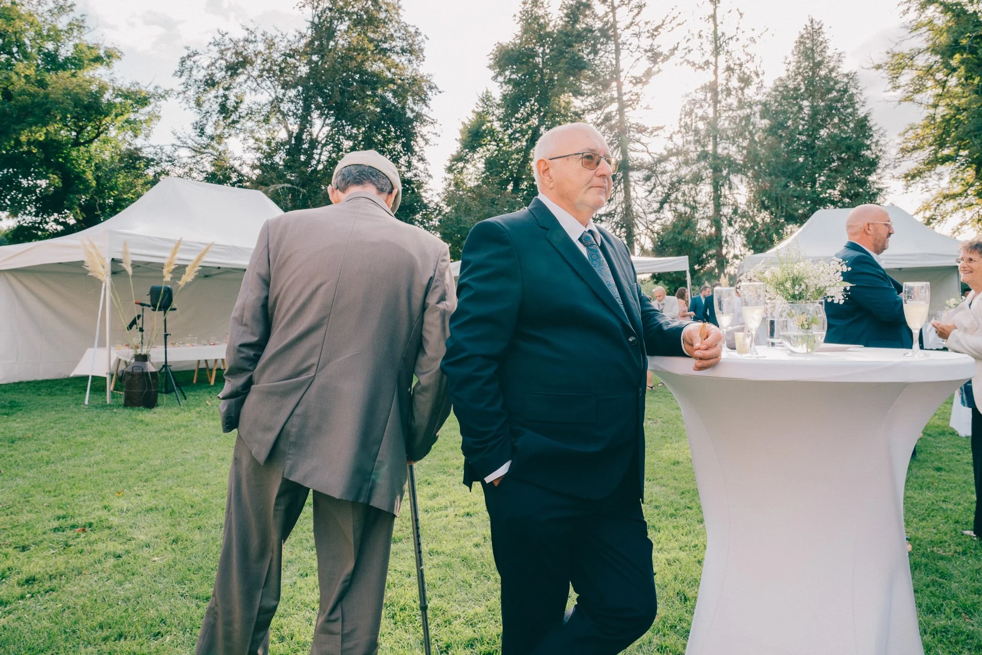 Gens lors d'un événement en plein air avec des tentes blanches, des tables avec des décorations florales et des personnes en tenue formelle, sous un ciel ensoleillé avec des arbres en arrière-plan.