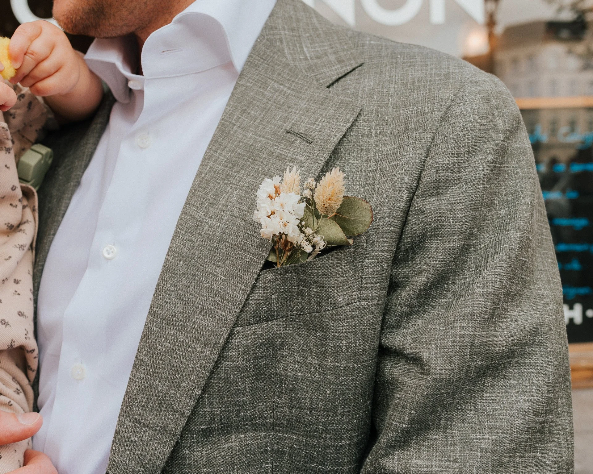 Homme portant un costume gris avec une boutonnière de fleurs dans la veste, mangeant quelque chose, en arrière-plan un bar ou une cave à vin.