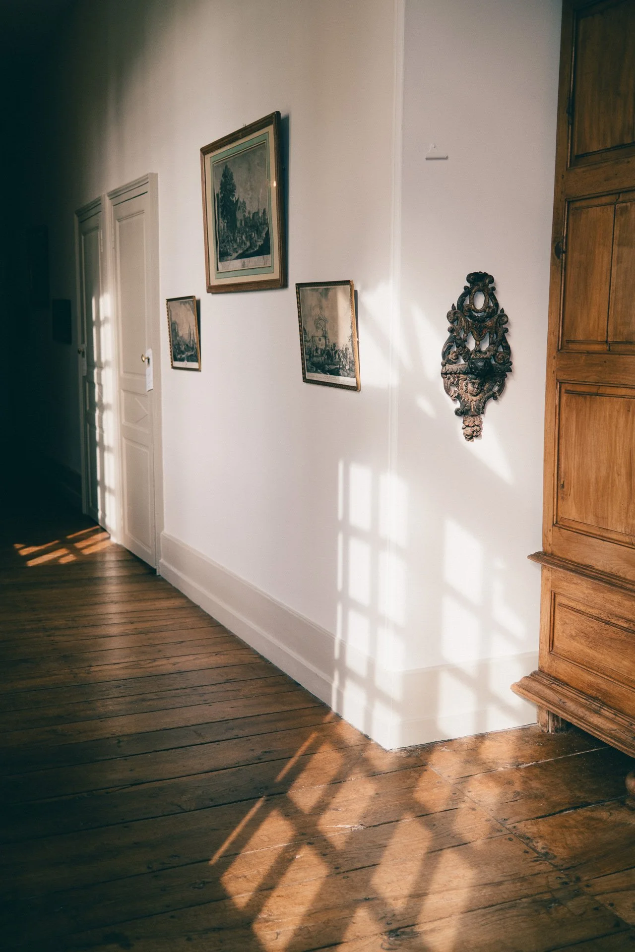 Un mur blanc dans une pièce avec plusieurs tableaux accrochés, une sculpture décorative en bois, et un meuble en bois massif. Le sol est en parquet en bois foncé, et la lumière naturelle crée des ombres sur le sol.