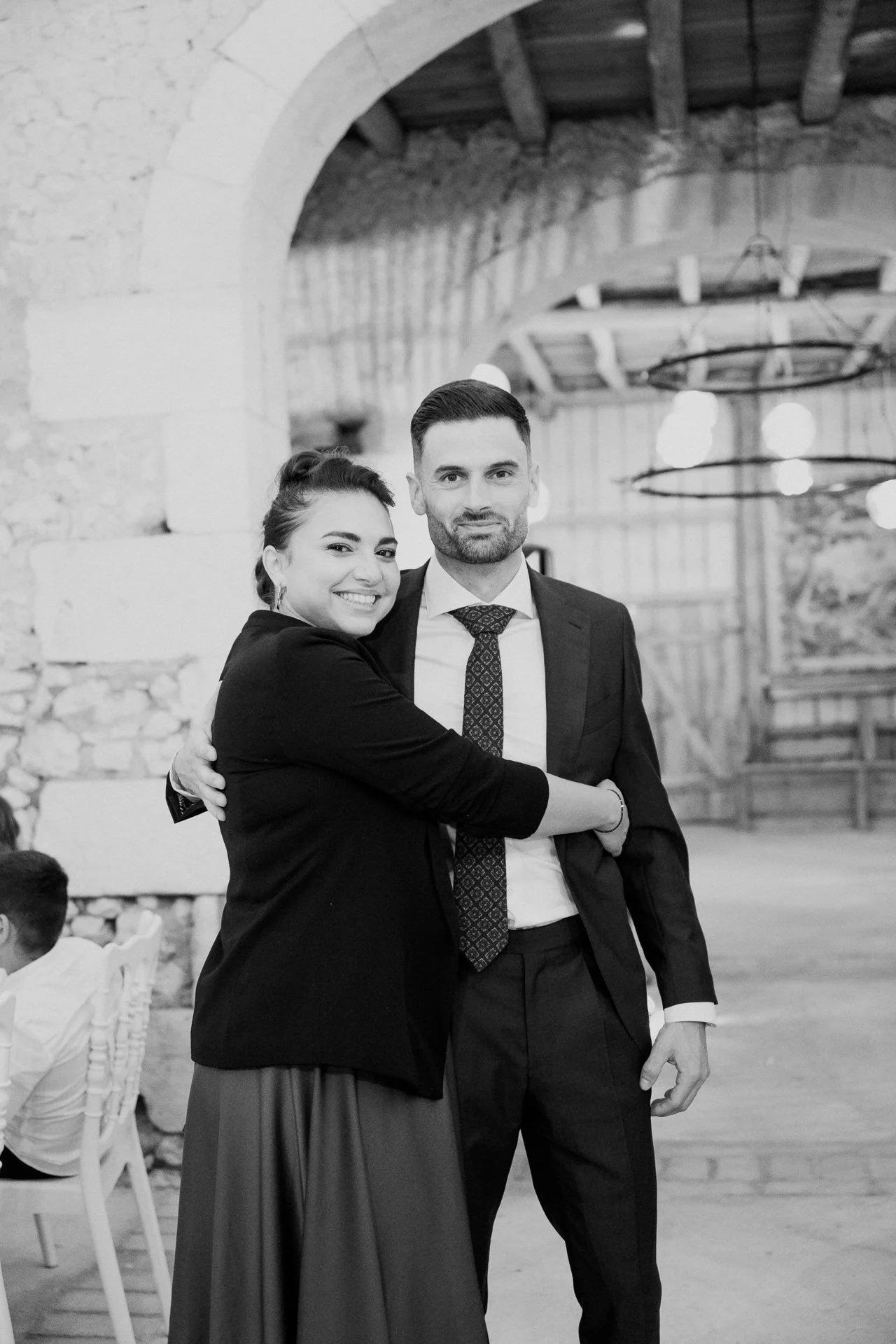 Un couple habillé élégamment, en train de danser ou de poser pour une photo dans un lieu intérieur rustique avec poutres en bois et murs en pierre.