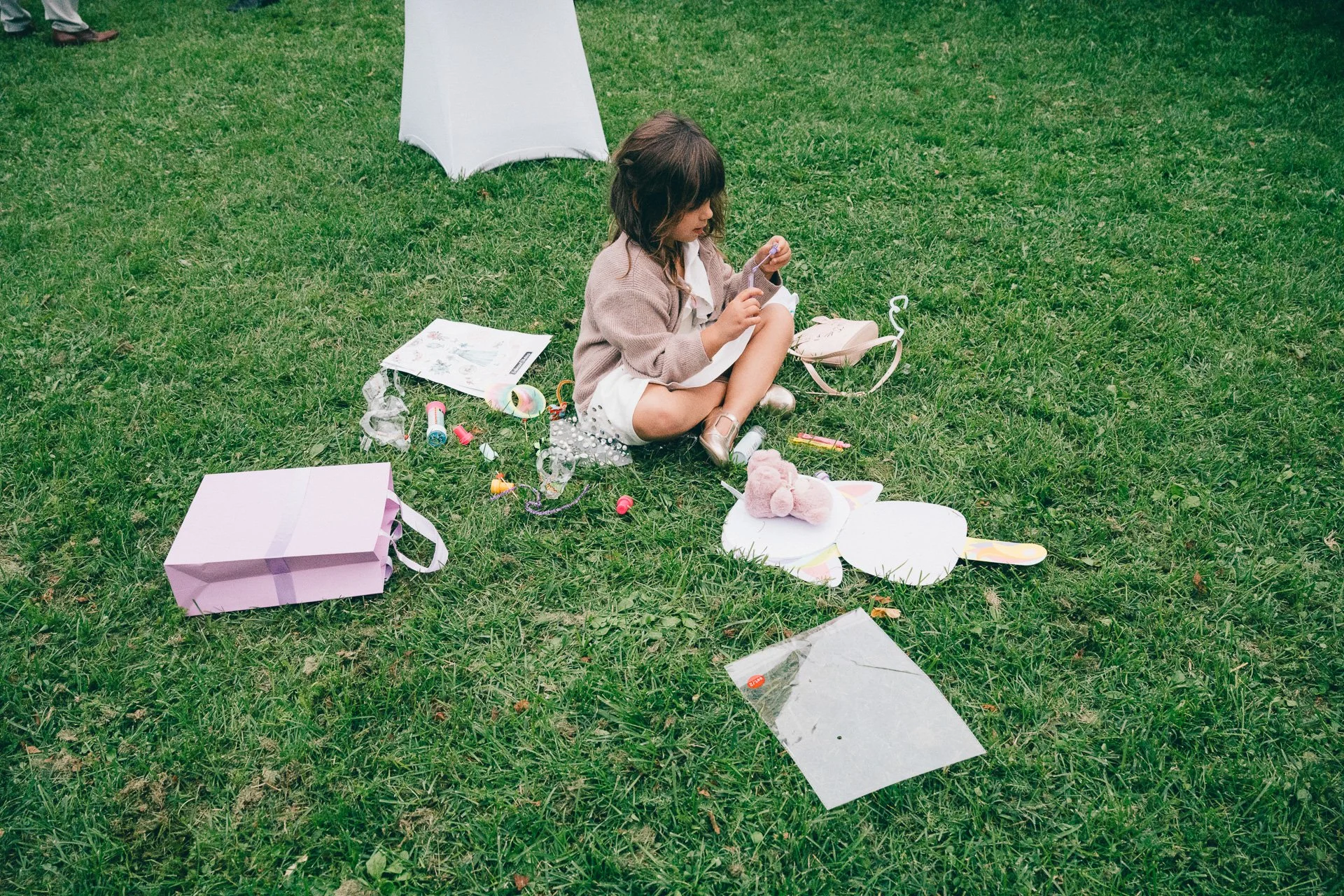 Une petite fille assise sur l'herbe, entourée de jouets, de papier et de boîte à surprises, lors d'une fête d'anniversaire en plein air.