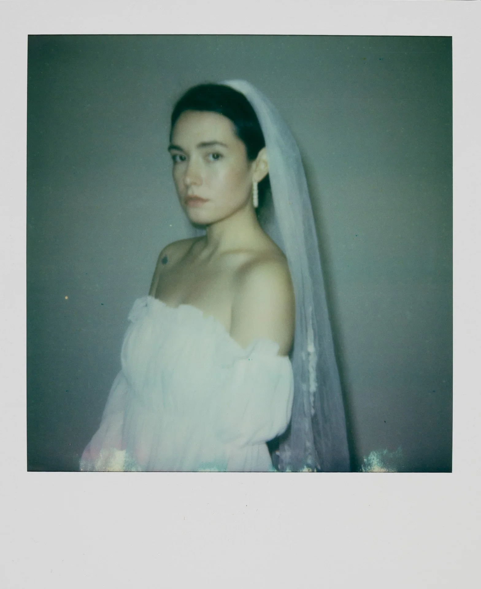 Un portrait d'une femme portant une robe blanche avec un voile blanc, portant des boucles d'oreilles longues, avec un fond neutre.