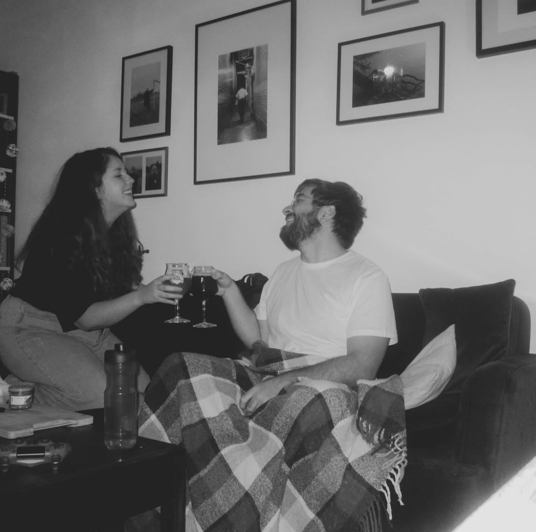Deux personnes, un homme et une femme, souriantes, tenant des verres, assises sur un canapé, dans un salon avec des cadres photos sur le mur.