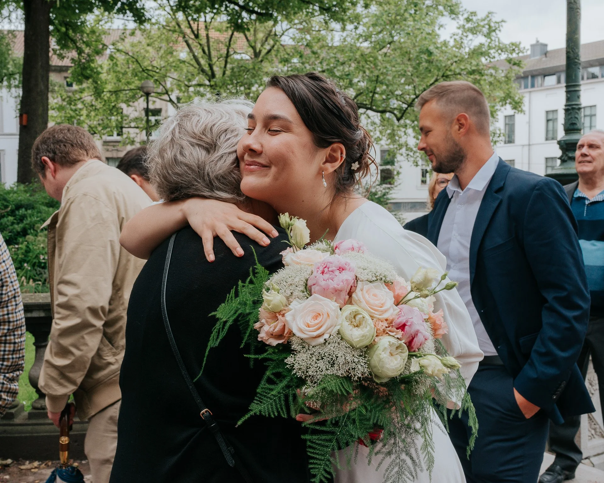 Une femme joyeuse embrasse une autre femme lors d'une fête, tenant un grand bouquet de fleurs roses et blanches, avec plusieurs personnes en arrière-plan dans un parc arboré.