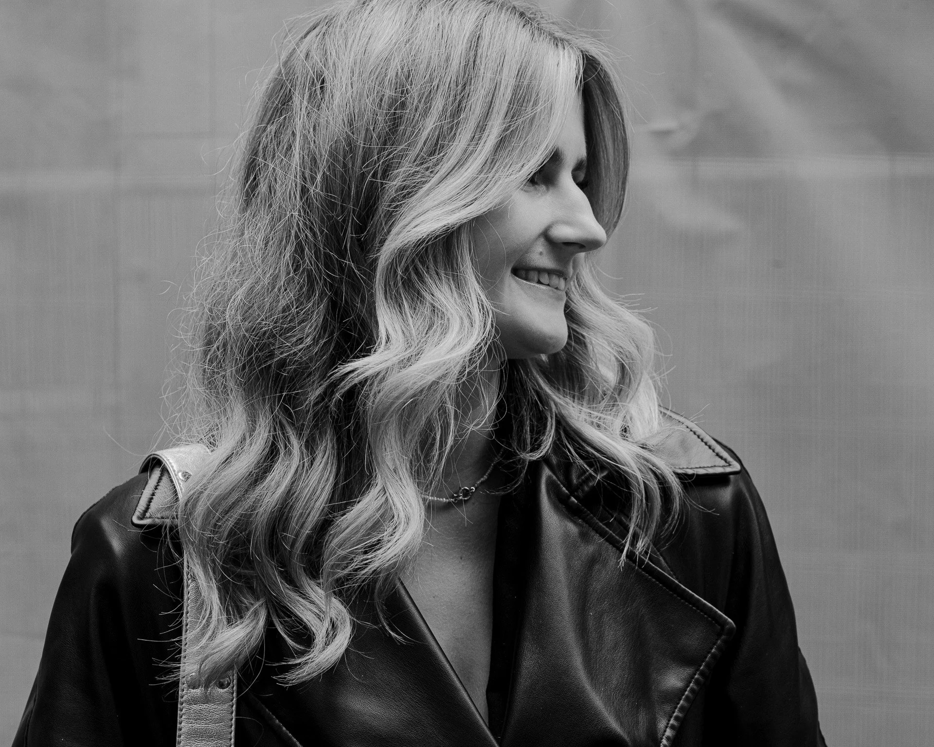 Portrait noir et blanc d'une femme souriante avec de longs cheveux bouclés portant une veste en cuir.
