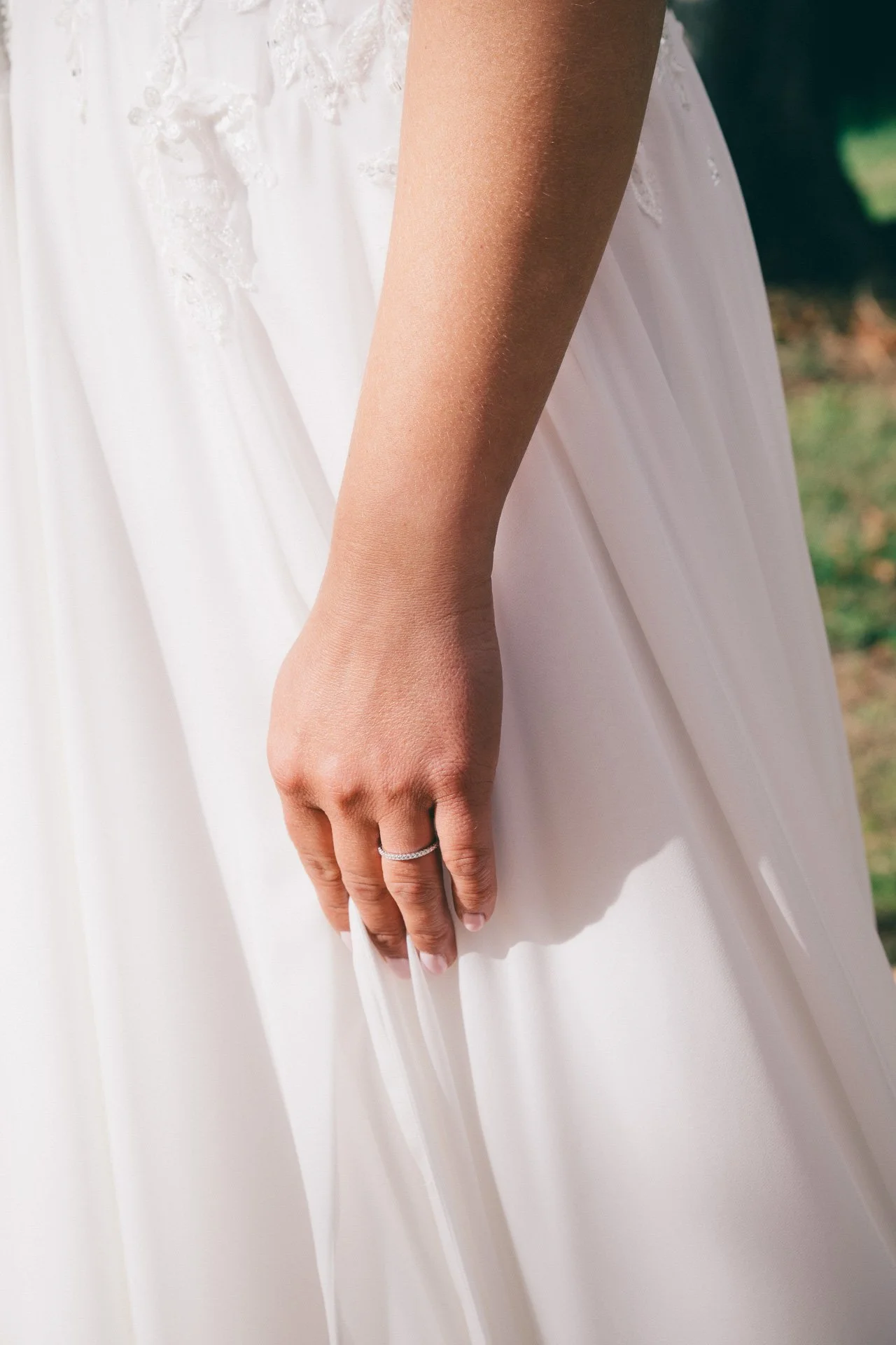 Une main féminine portant une alliance, tenant un tissu blanc devant une robe de mariage. La main est nue, avec un seul anneau en argent ou en diamant. L'arrière-plan est flou, avec de la verdure visible.
