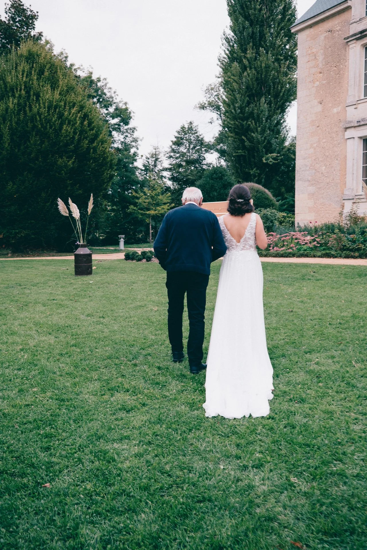 Deux personnes, un homme âgé et une femme en robe de mariage, marchent dans un jardin vert avec des arbres et un bâtiment ancien en pierre, dans une ambiance de mariage ou de cérémonie.