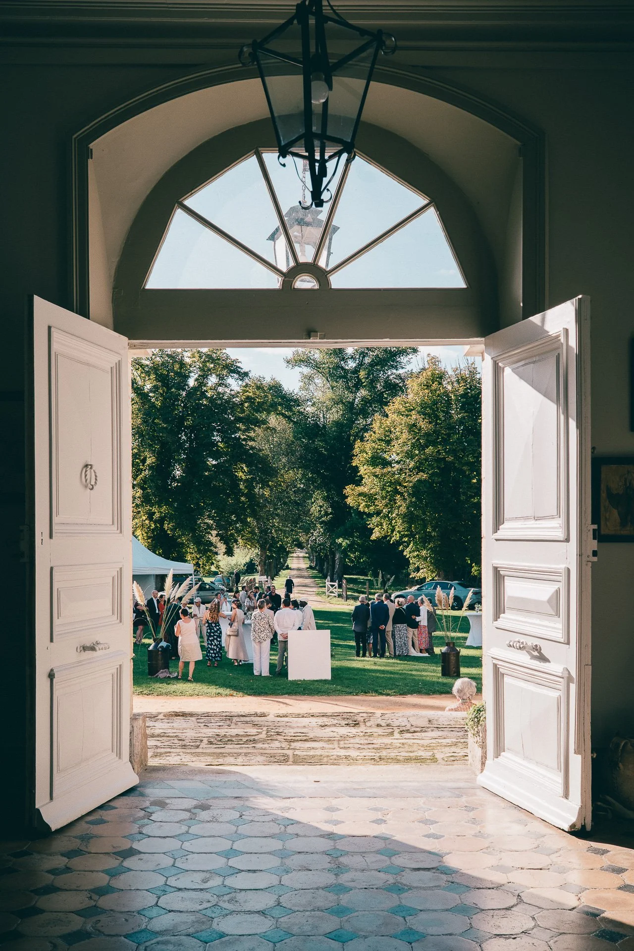 Vue à travers une grande porte en bois blanc ouverte qui mène à un événement en plein air avec des personnes réunies sur une pelouse verte, entourée d'arbres, lors d'une journée ensoleillée.