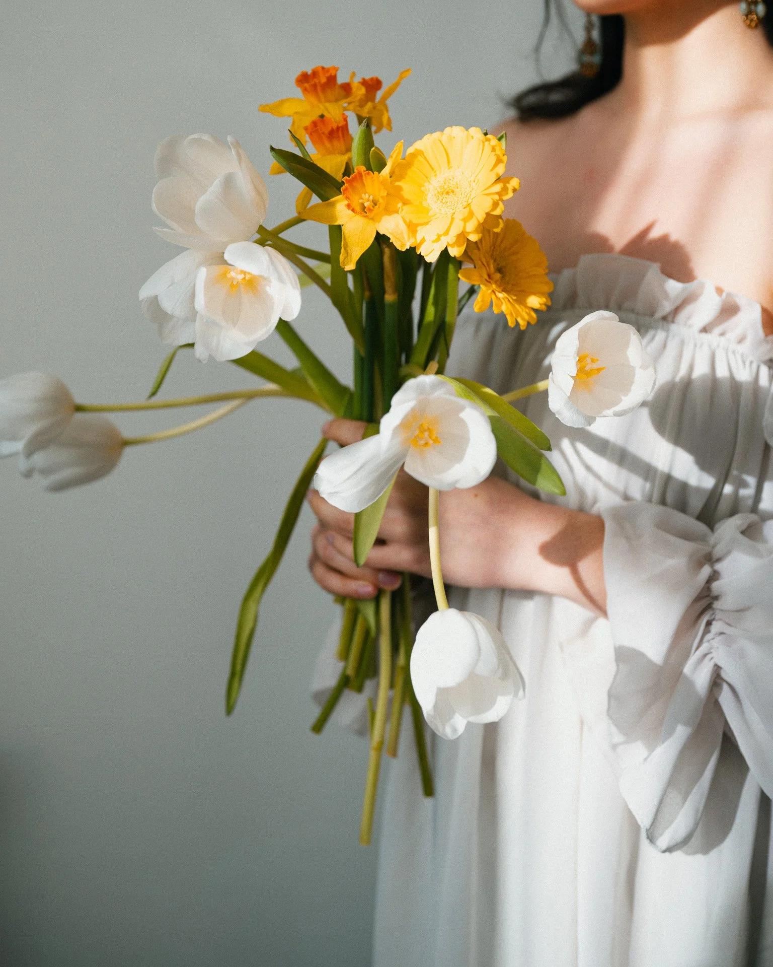 Femme tenant un bouquet de fleurs blanches, jaunes et orange dans une robe blanche à manches longues. La photo est prise en gros plan, montrant surtout le bouquet et une partie de la robe.