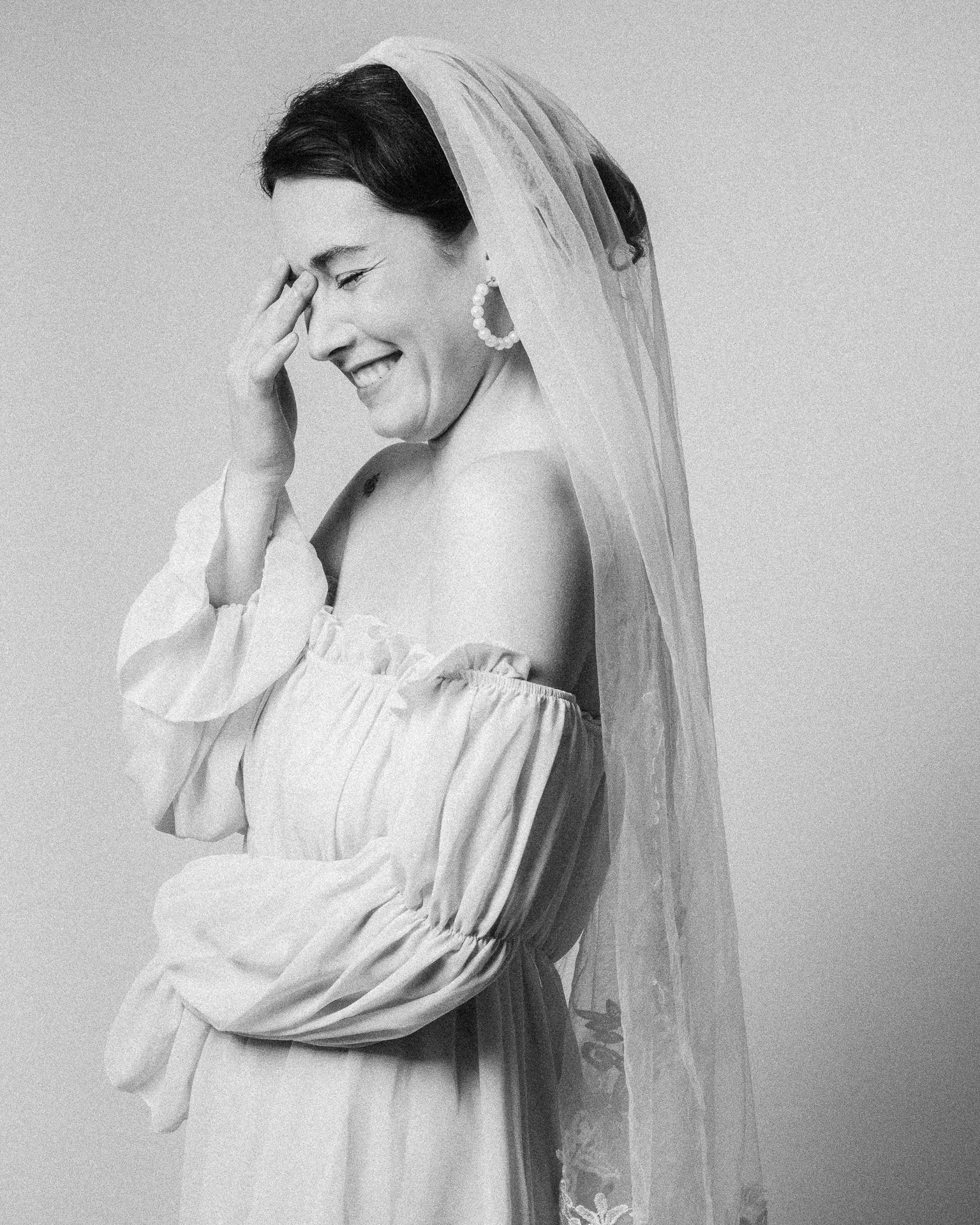 Une femme souriante porte une robe blanche et un voile de mariée, avec une boucle d'oreille en perles, la main sur le front, en noir et blanc.