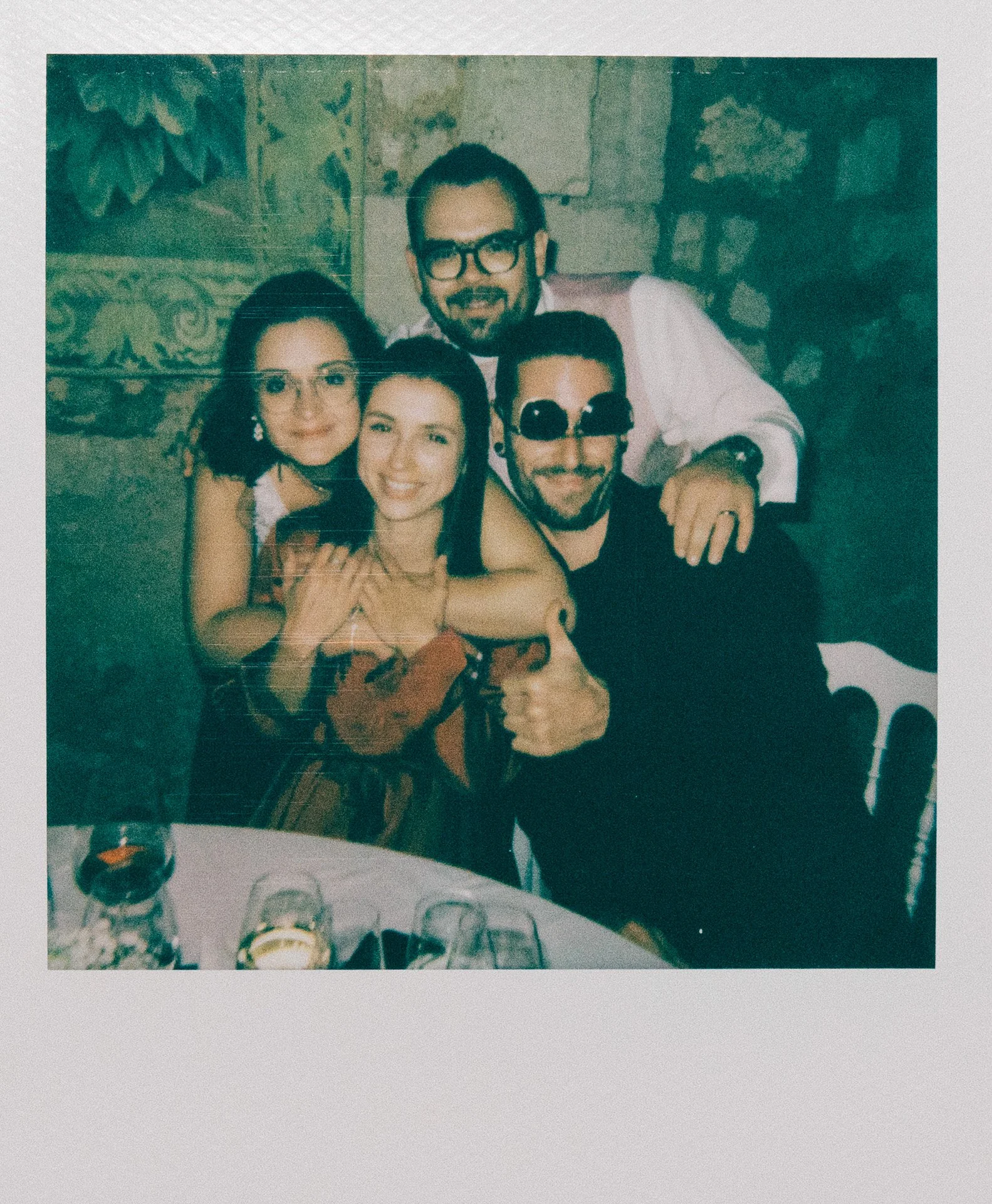 Groupe de cinq amis souriants lors d'une fête, assis autour d'une table avec des verres et une bouteille. Ambiance festive avec décorations en arrière-plan.