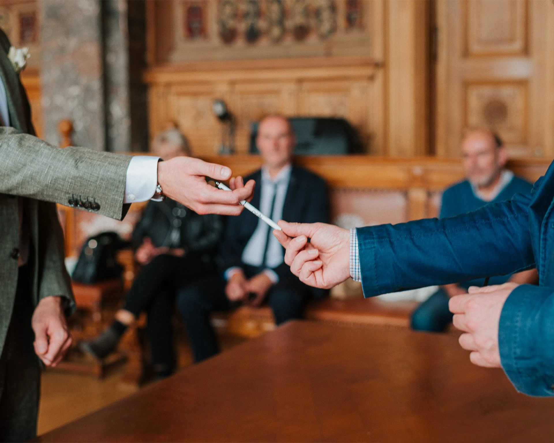 Une personne en costume remet un stylo à une autre personne dans une salle en bois, avec des hommes en arrière-plan assis et regardant la scène.