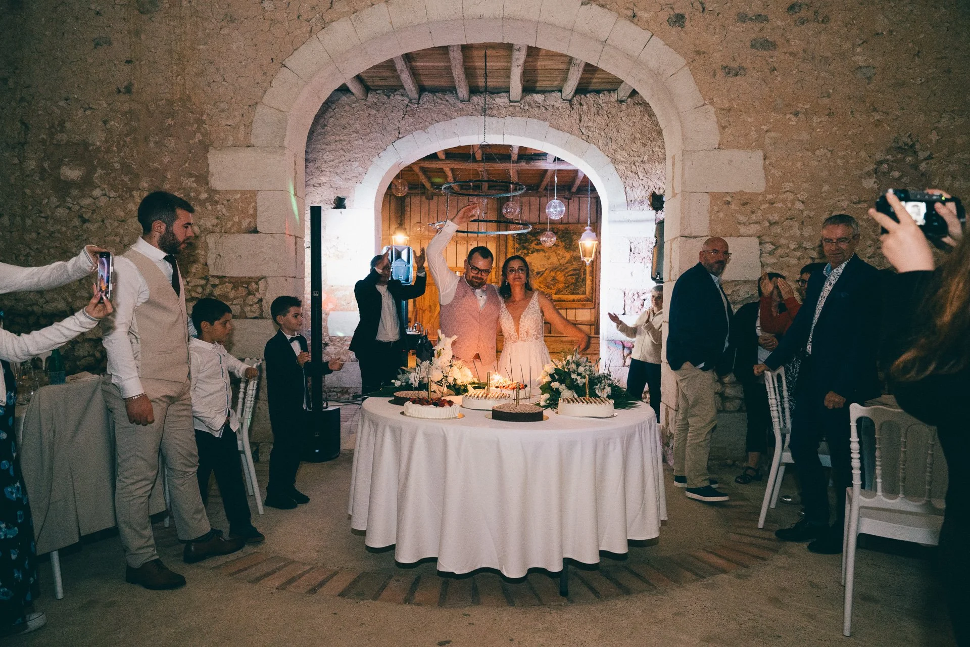 Une célébration de mariage où le marié et la mariée se tiennent derrière un gâteau au centre de la table, entourés d'invités. La scène se déroule dans un lieu avec des murs en pierre et un plafond en bois, décoré avec des lampes suspendues et une pei