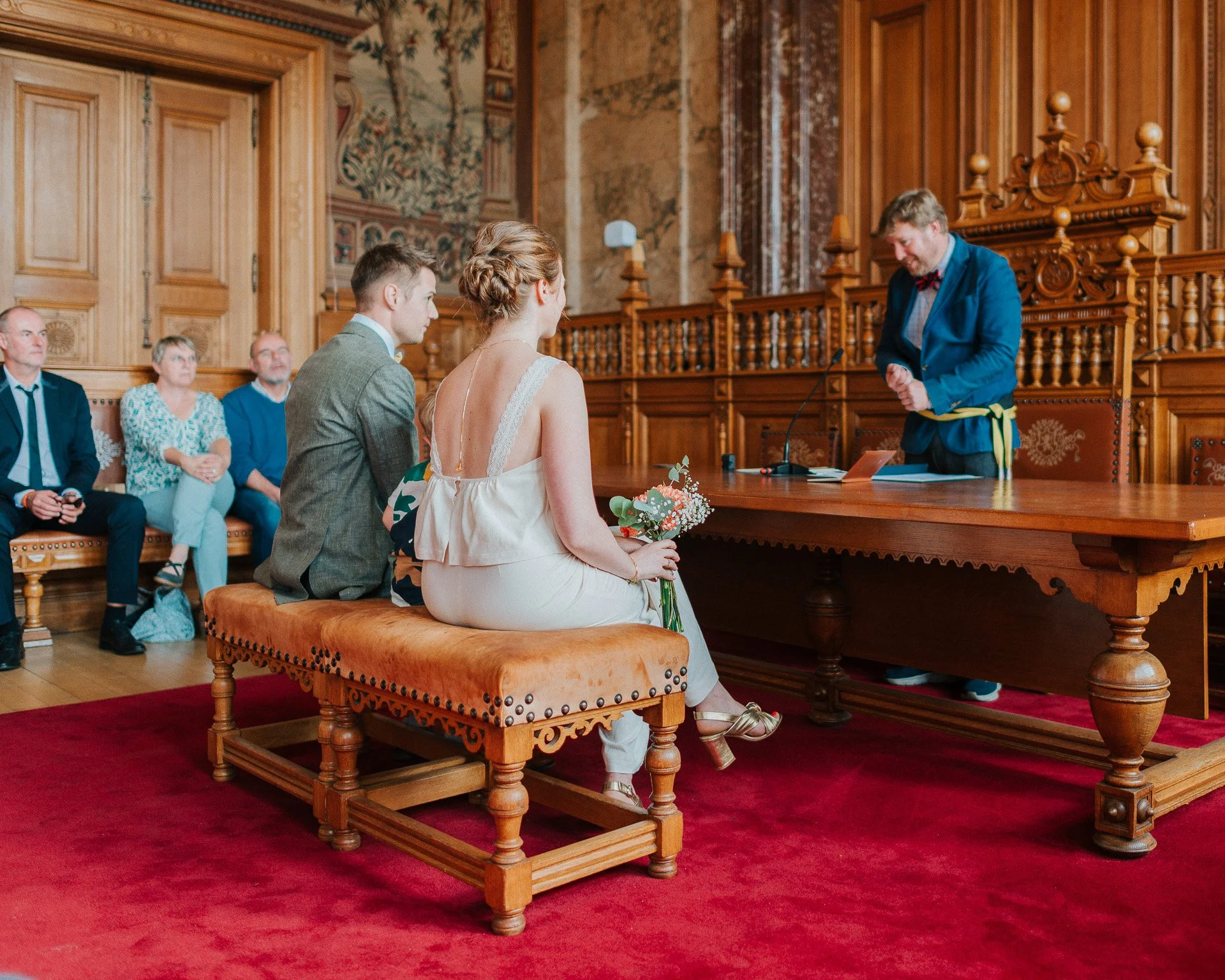 Un couple lors d'une cérémonie nuptiale assis sur un banc en bois dans une salle ornée en bois sculpté et tapis rouge, un officier célèbre le mariage.