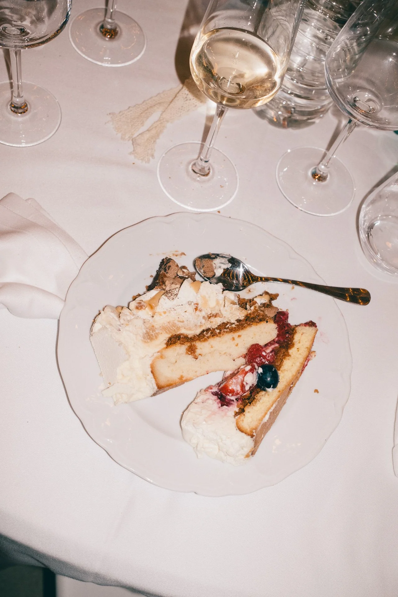 Une part de gâteau aux fruits rouges sur une assiette, accompagnée d'une cuillère, sur une table avec une nappe blanche, plusieurs verres en arrière-plan, dont certains remplis de vin blanc et d'eau.