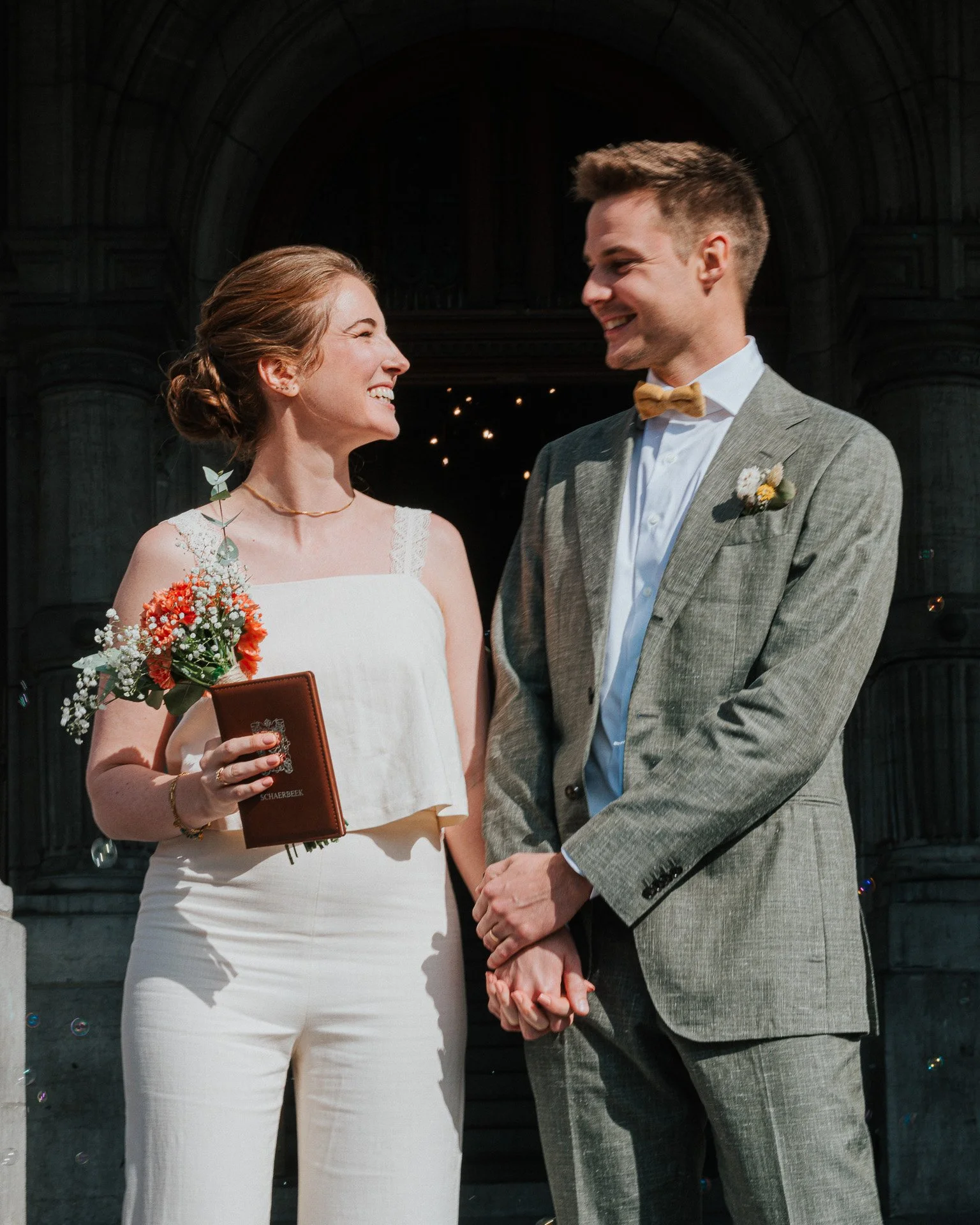 Un couple lors d'une cérémonie de mariage dans une église, la femme en robe blanche avec un bouquet de fleurs rouges et blanches, l'homme en costume gris avec un nœud papillon beige, se tenant la main et souriant à l'autel.